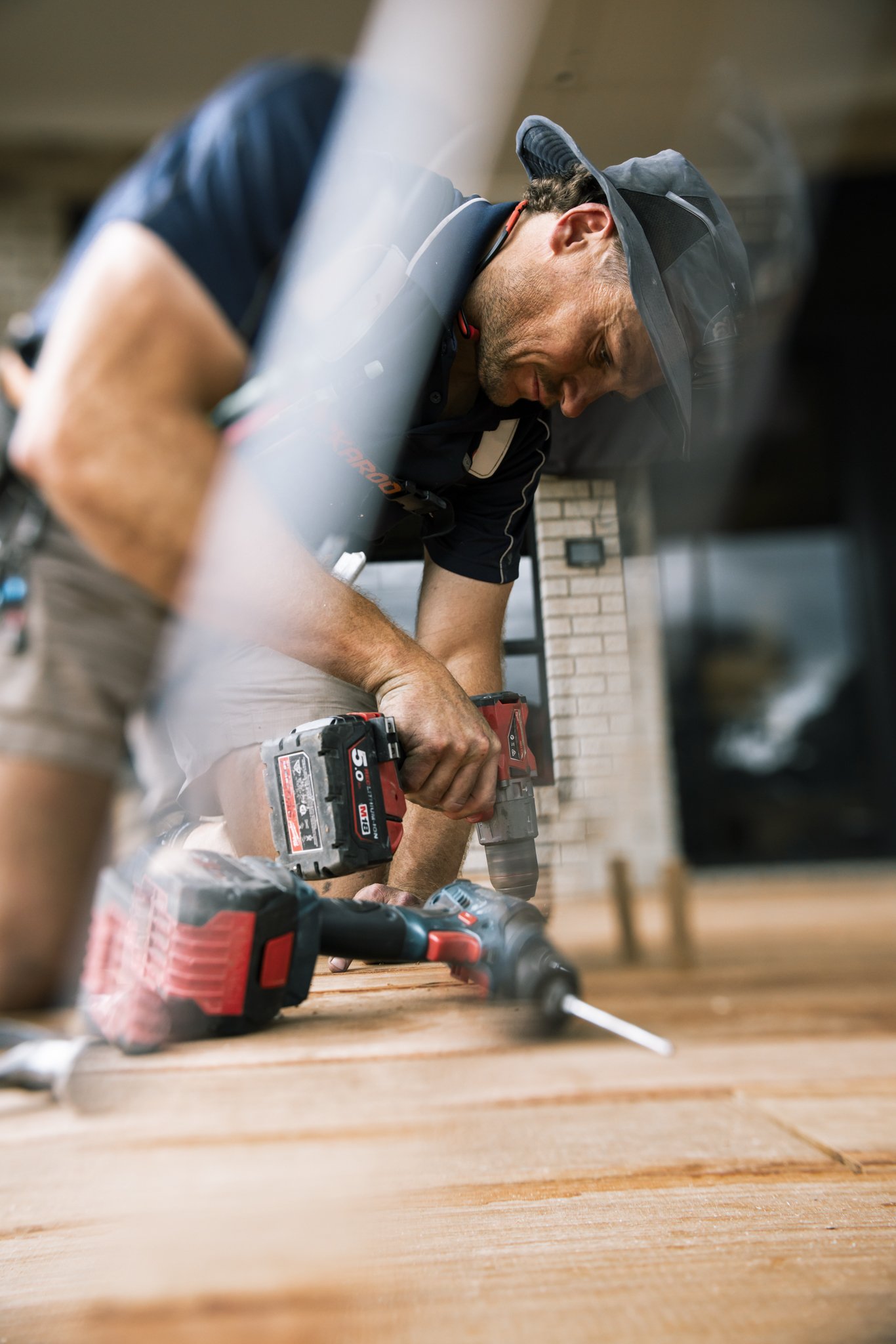 Man working on hardwood floor with power drill, wearing a cap, navy shirt, and beige shorts, indoors with a brick fireplace in the background.
