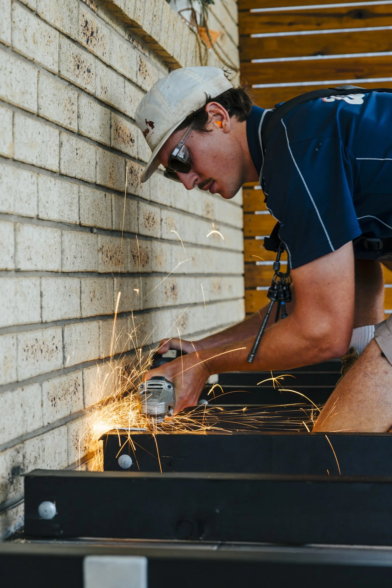 Man wearing work goggles and a hat, using a grinder to cut or grind metal on a black surface outdoors against a brick wall with a wooden fence in the background.