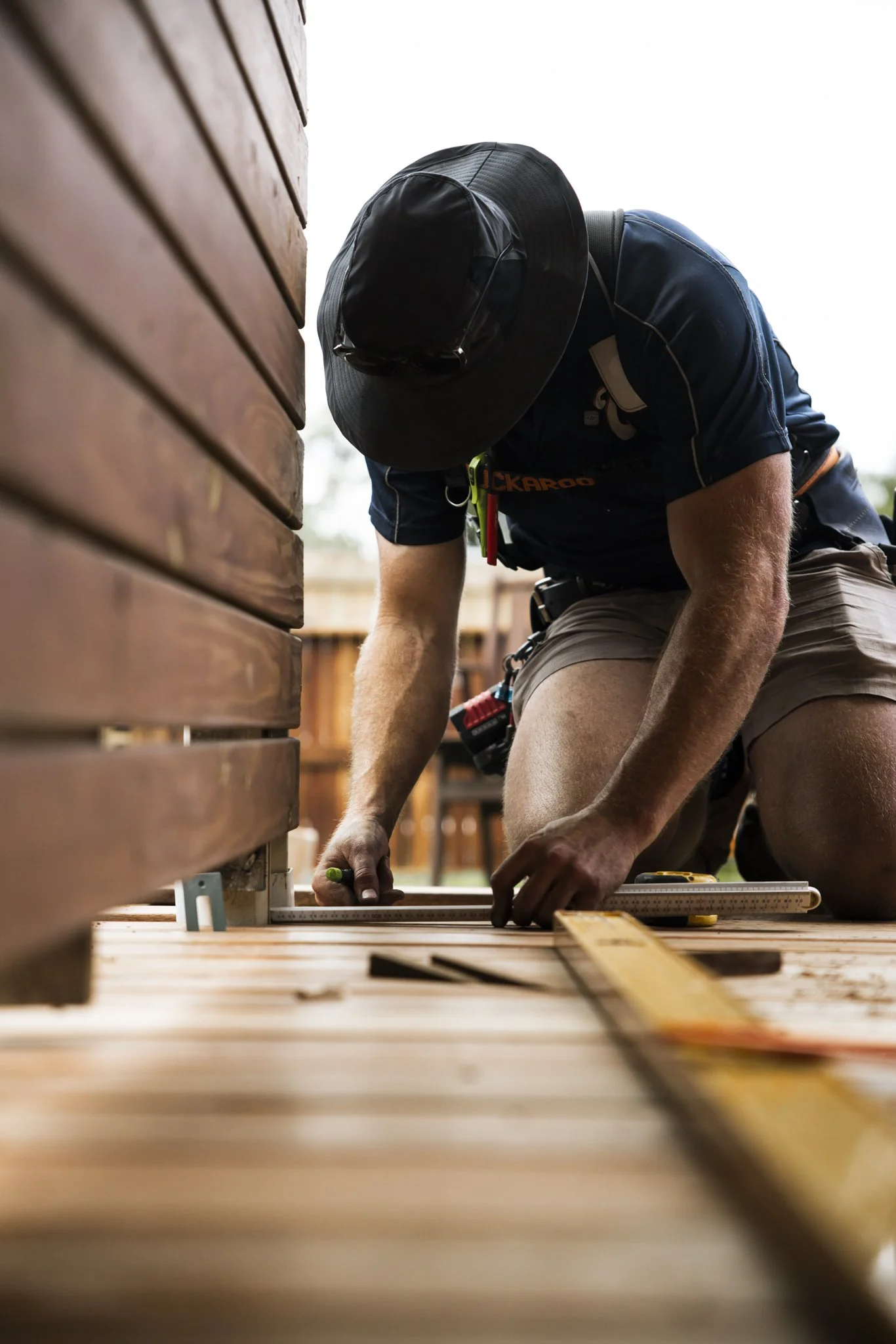 A person wearing a wide-brimmed hat and outdoor work attire is kneeling on a wooden deck, measuring and marking the wood with a square and a measuring tape, surrounded by tools.