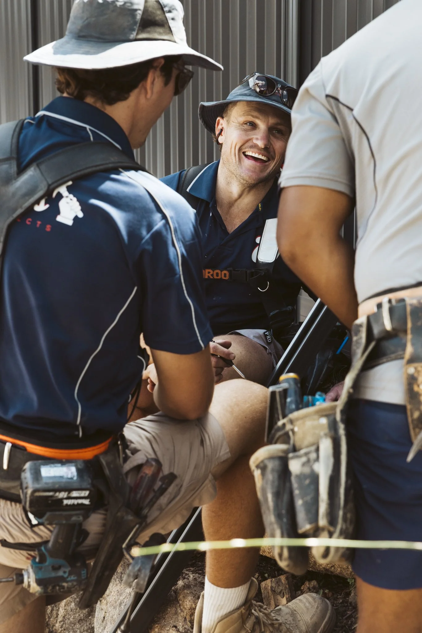 Three men, two wearing wide-brimmed hats and one with sunglasses, are gathered around an injured person sitting on a chair outdoors, with a gray metal wall behind them. They appear to be engaged in providing emergency assistance.
