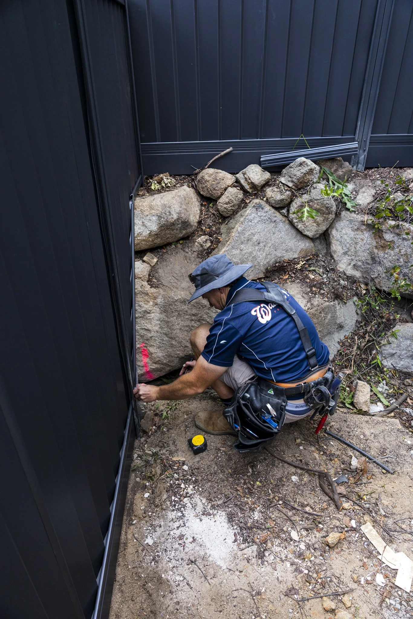 A man installing a black metal fence or wall outdoors, kneeling on the dirt with tools and measuring tape, next to rock landscaping.