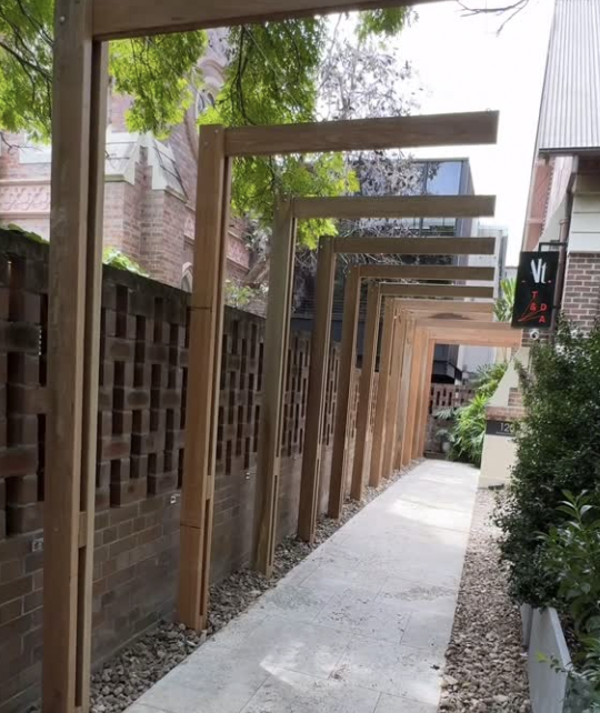 A sidewalk with wooden pergolas extending along a brick wall, with green trees and bushes nearby.