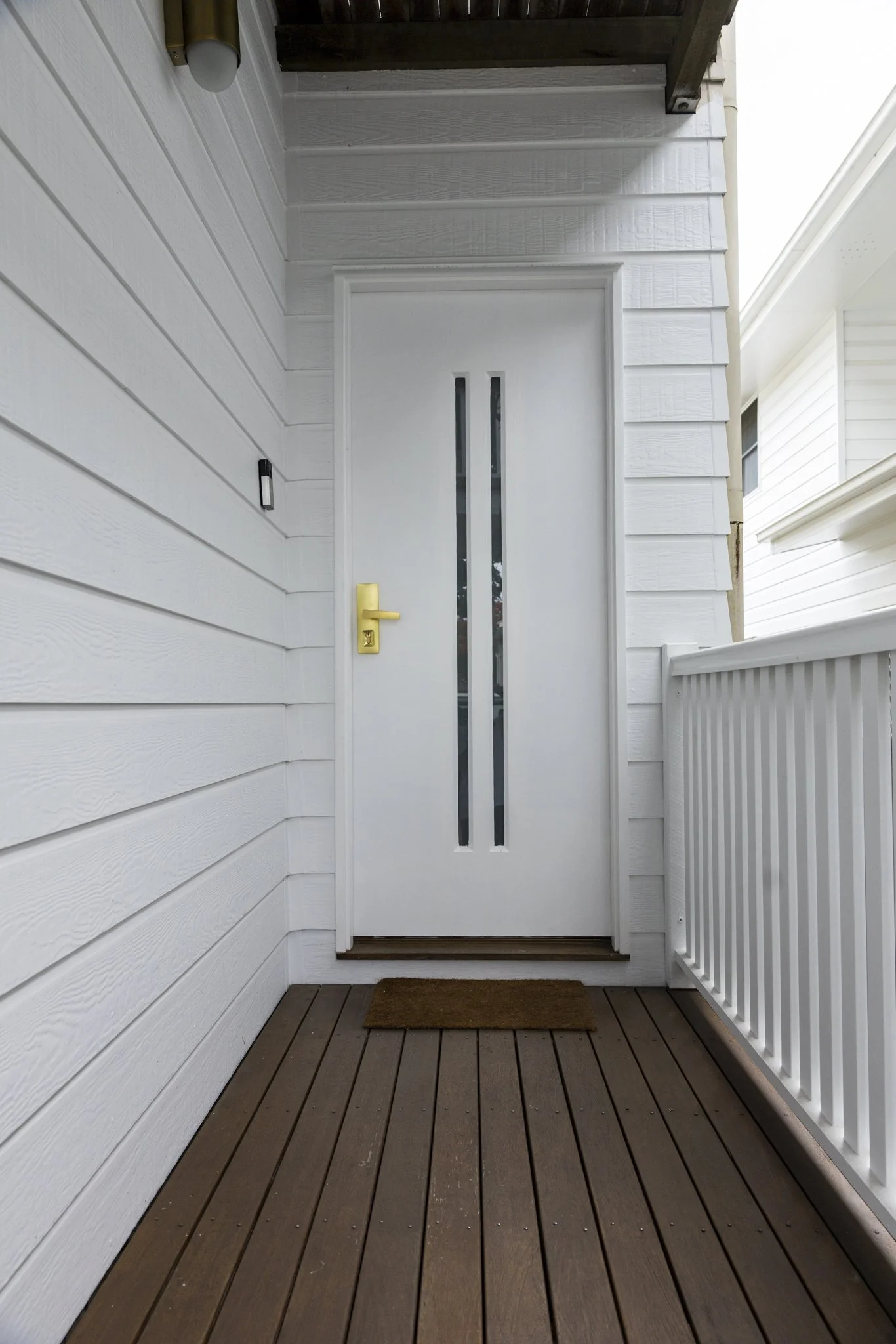Front door with vertical glass panels, white exterior wall with horizontal siding, wood porch flooring, and a small brown welcome mat.