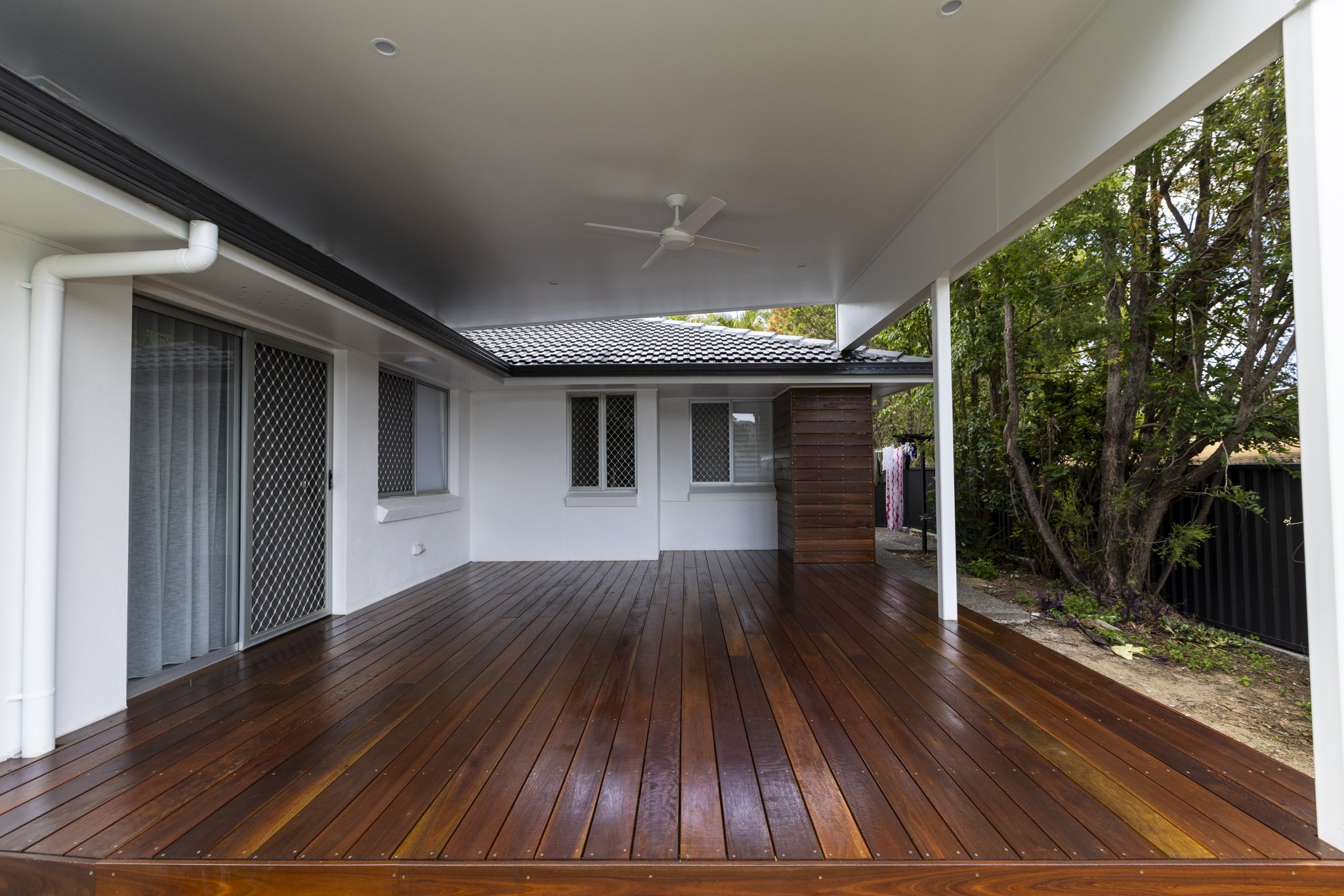 Modern backyard with polished wooden deck, white house exterior, sliding glass door, windows with security screens, and a ceiling fan under covered patio, surrounded by lush trees.