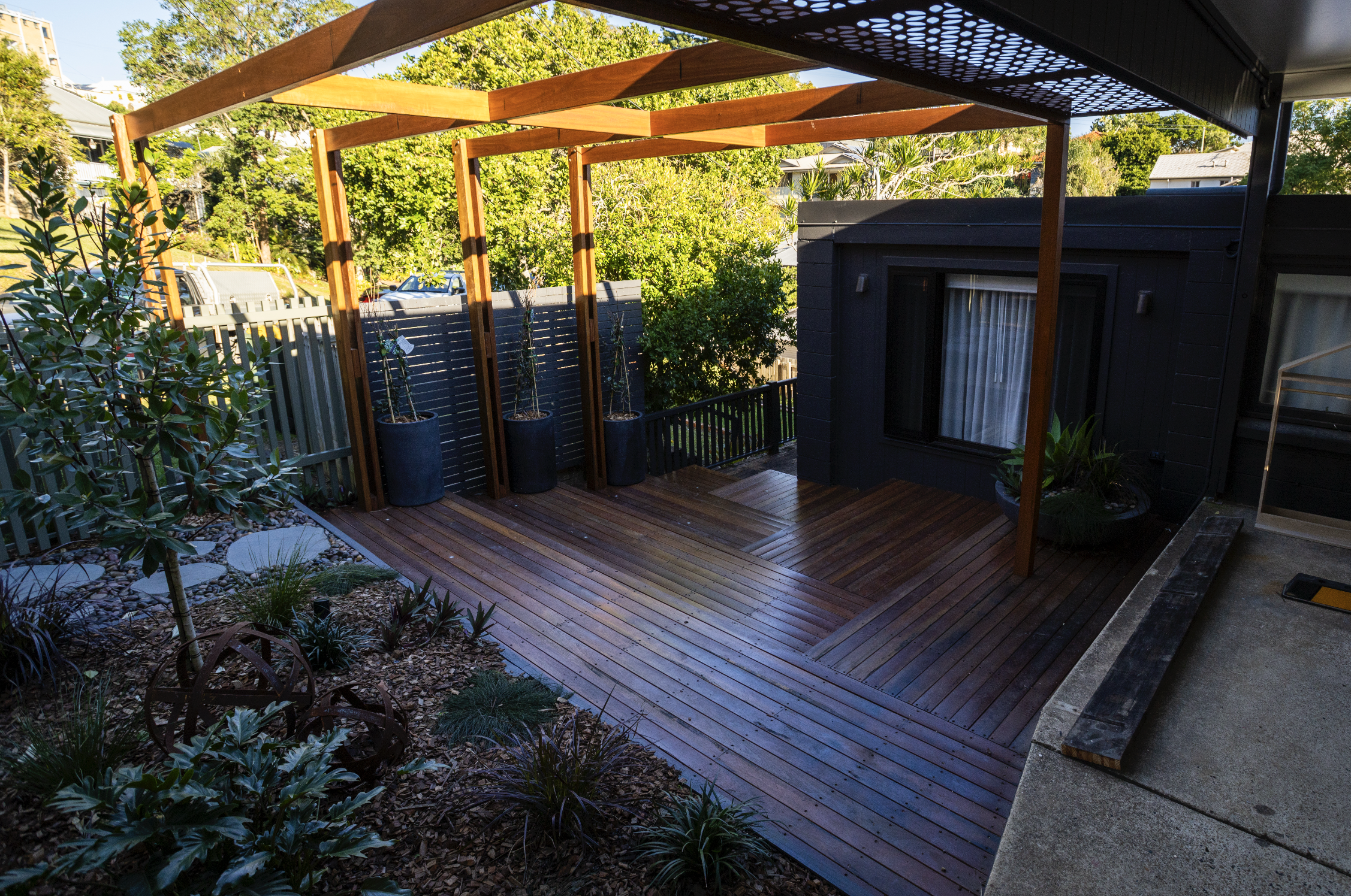Two construction workers building a wooden deck in a backyard. They are surrounded by trees and a white fence. The partially completed deck is visible, with wooden planks being laid out. The workers are wearing blue shirts, shorts, and hats.
