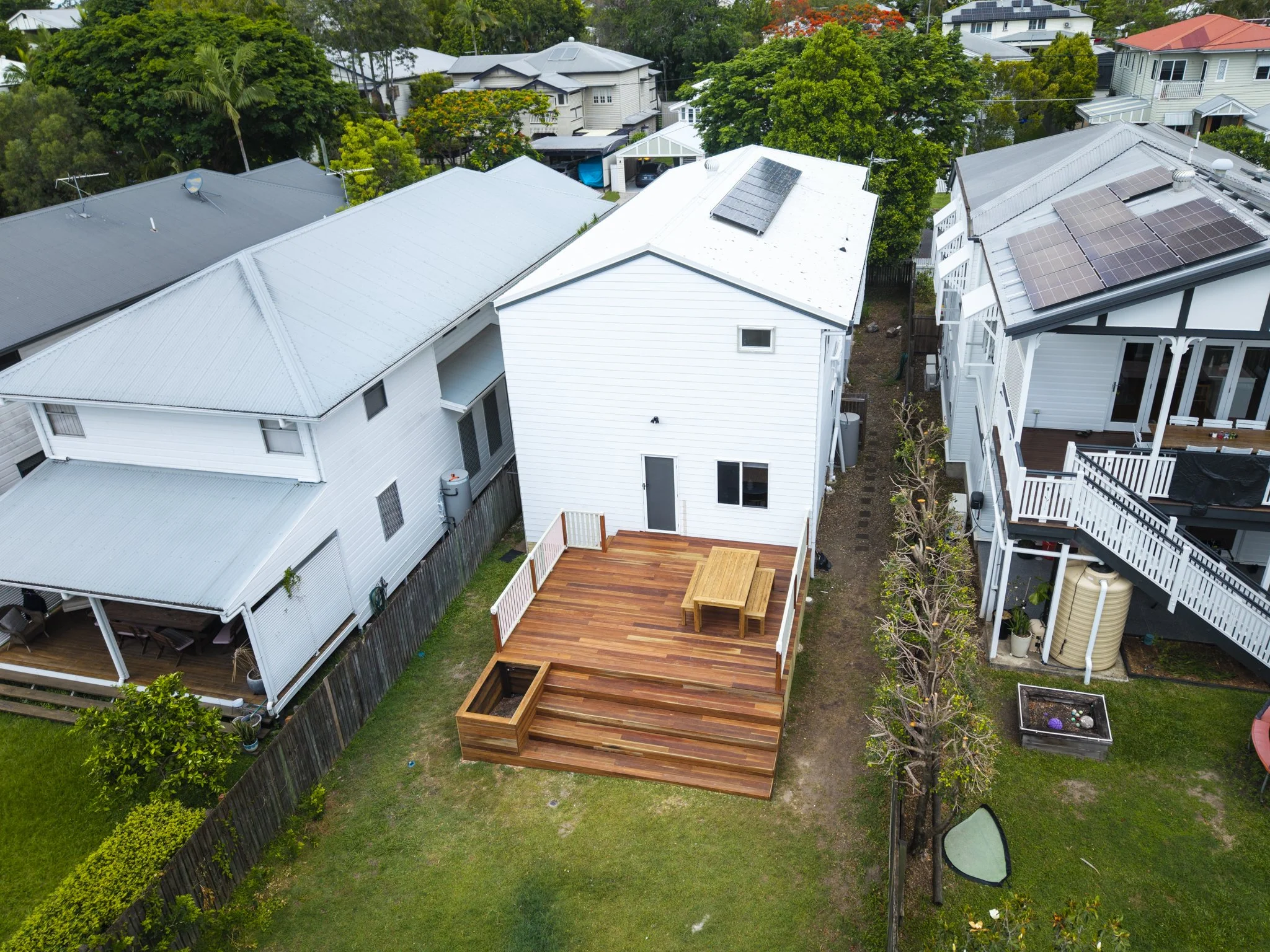 Aerial view of a backyard with a wooden deck featuring a built-in fire pit, a picnic table, and surrounding grass, adjacent to a white house with solar panels on the roof, neighboring houses, trees, and a clear sky.