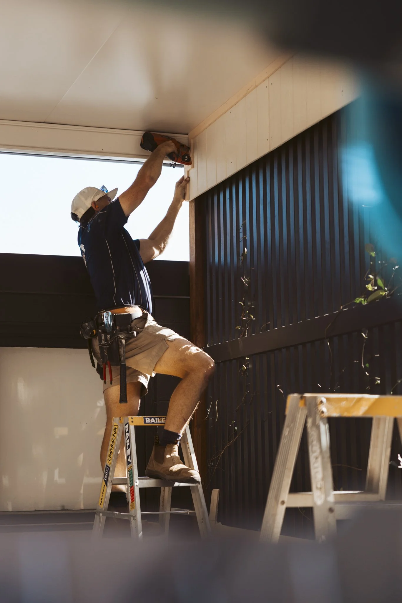 Man standing on a ladder installing or repairing a ceiling with a cordless drill, wearing a cap, shorts, and a tool belt.