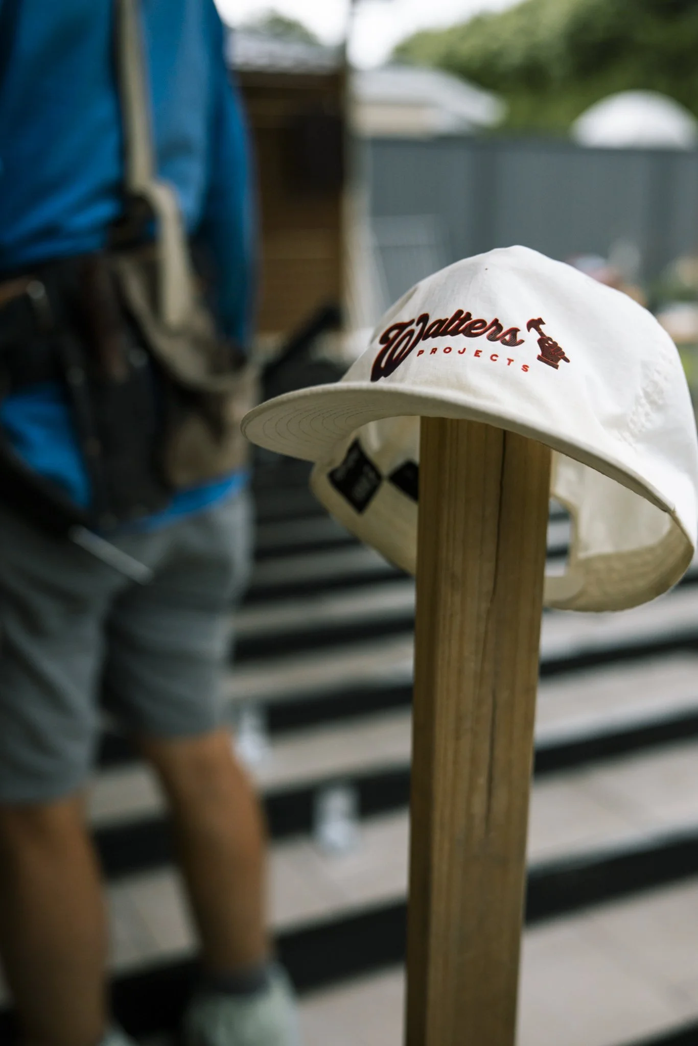 A white hat with the words 'Valters Projects' embroidered in red on the front, mounted on a wooden post, with a person in a blue shirt and gray shorts blurred in the background.