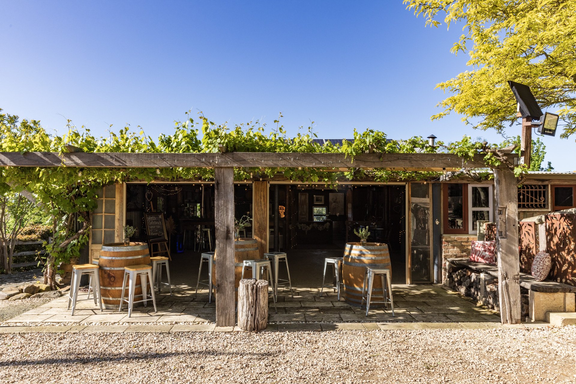 Exterior view of rustic outdoor bar and seating area with grapevine-covered pergola, metal stools, barrel tables, and wooden benches on stone patio, under a clear blue sky.