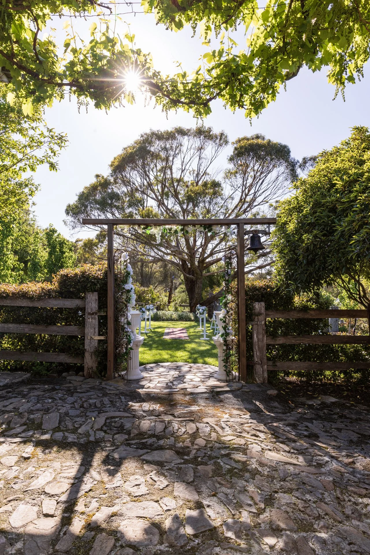 an outdoor wedding ceremony setup with a stone pathway leading to an arch decorated with flowers, in a lush green garden with trees and bushes, under a bright sunny sky