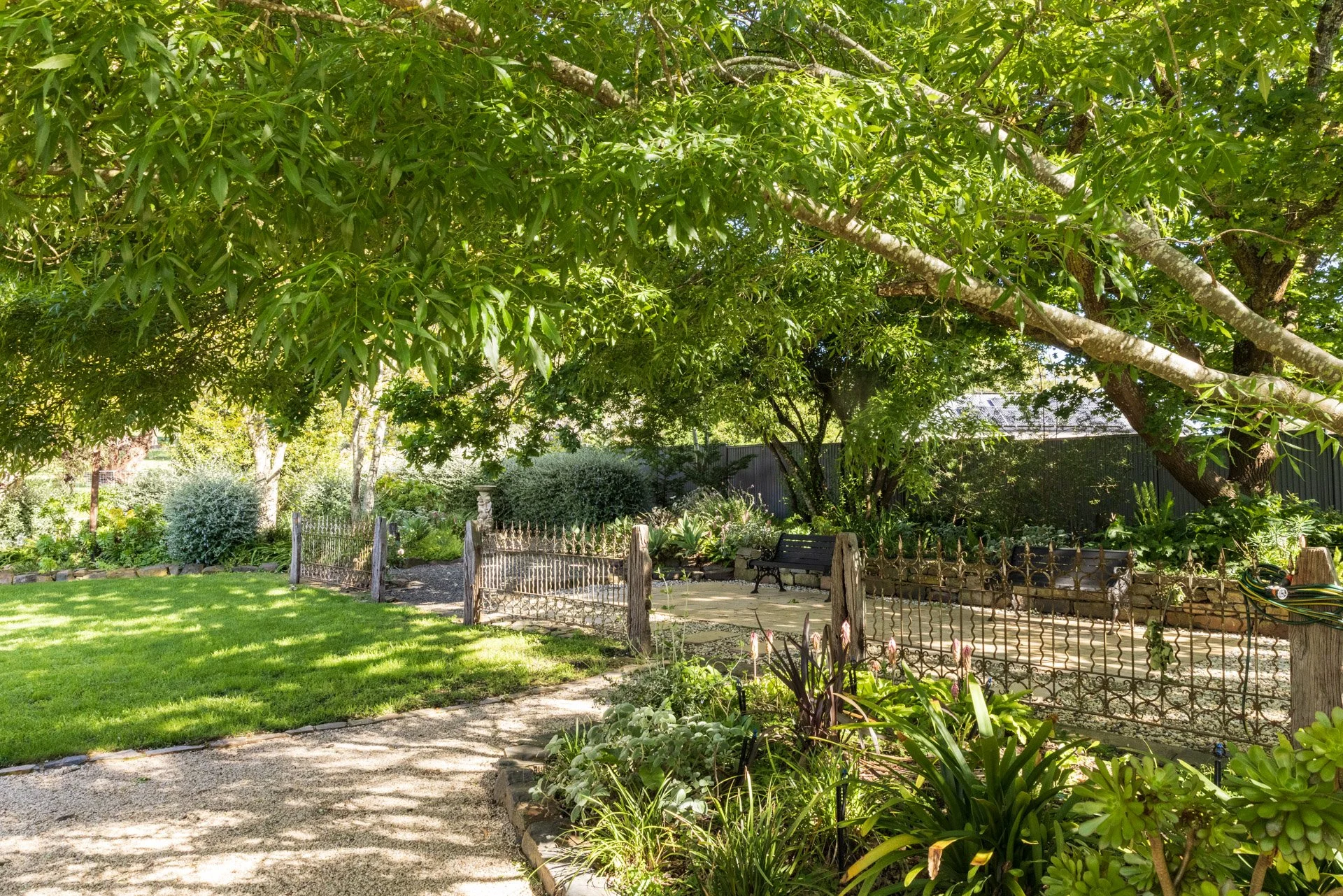 A lush backyard garden with a large tree providing shade, a small gravel pathway, a green lawn, and a decorative metal fence surrounding a patio area with benches and various plants.