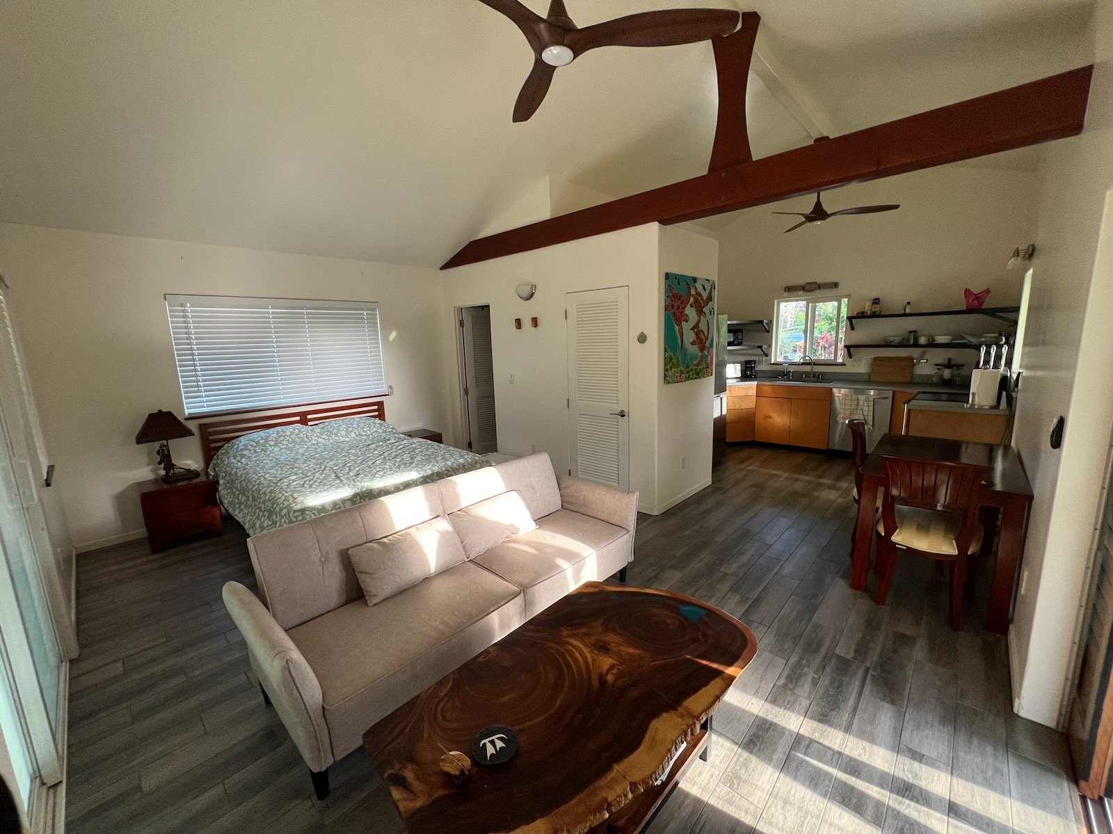 Open-plan living space with a bed, sofa, wooden table, and kitchen area featuring a dining table and appliances, under a vaulted ceiling with ceiling fans.