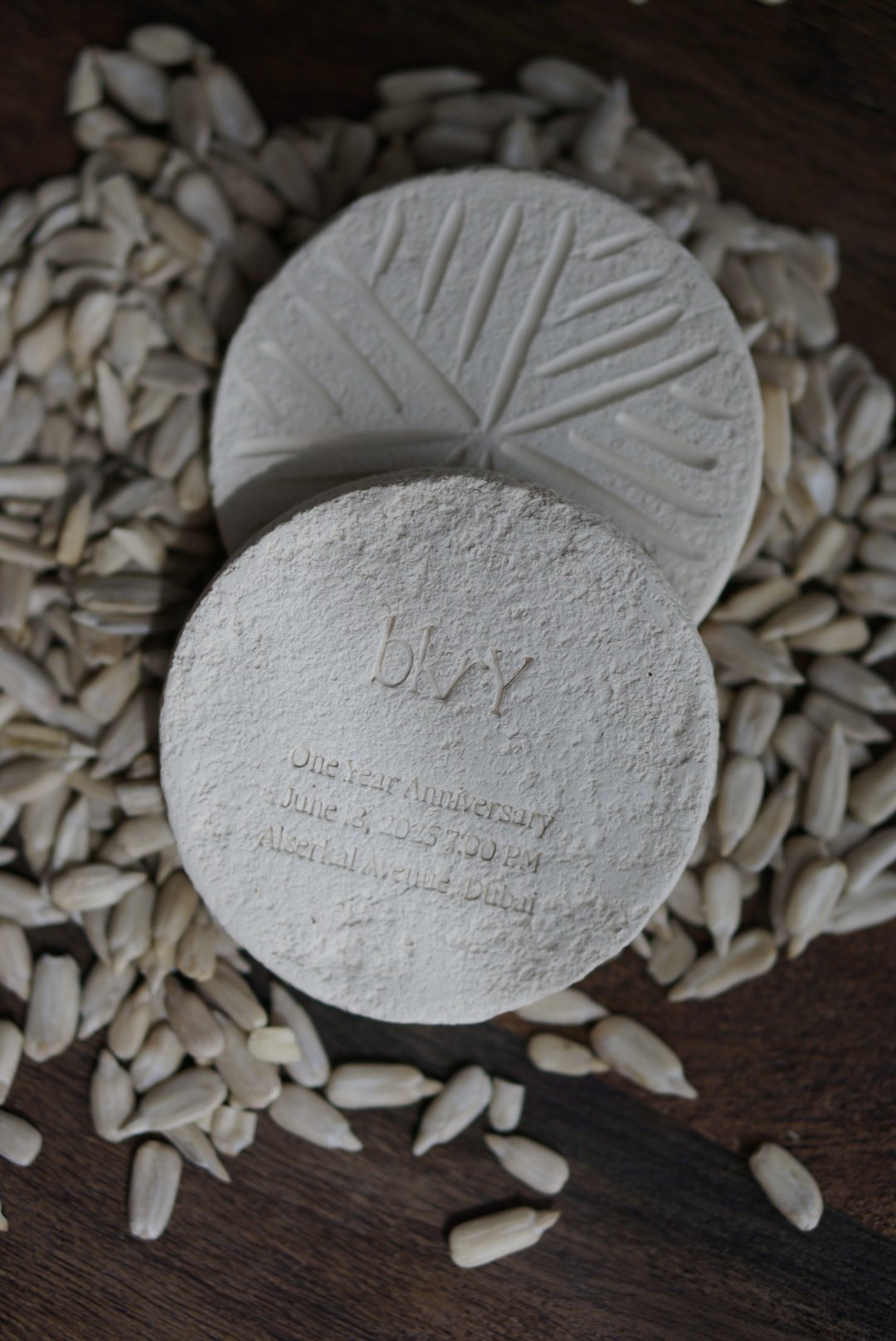 Two round, textured soaps on a wooden surface surrounded by sunflower seeds. One soap is embossed with the word 'by' and has an engraved message below, reading 'One-Year Anniversary June 18, 2015 7:00 PM Alserkal Avenue Dubai.' The other soap has a c