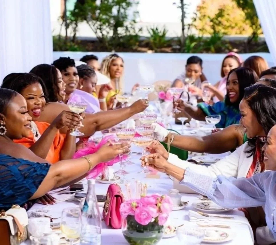A diverse group of women gathered at a long table outdoors, raising glasses in a toast during a celebration or special event. Clean Beauty Coalition