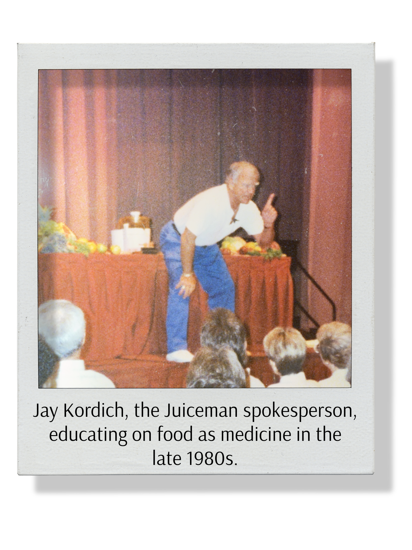 Photograph of Jay Kordich, the Juiceman, speaking at a presentation about food as medicine in the late 1980s, with fruit and vegetables on a table behind him, in front of an auditorium audience.