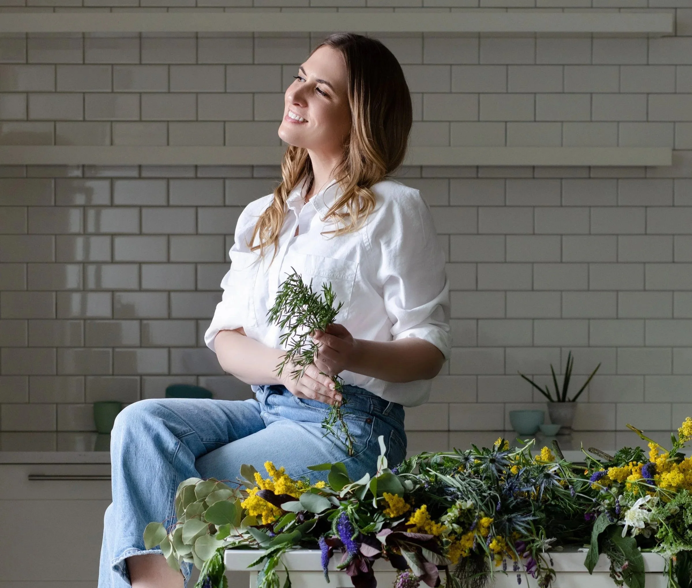 A woman with long, wavy brown hair wearing a white shirt and blue jeans, sitting indoors holding a small bouquet of green foliage. There are various flowers in front of her and a background of light-colored brick wall.