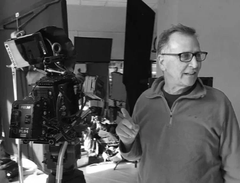 Man with glasses and a jacket standing next to a professional camera setup in a studio or production environment.