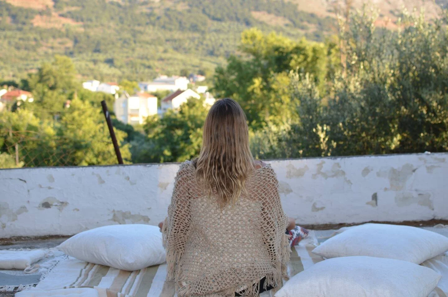 Dr. Michele Burklund  with long, curly blonde hair sitting on an outdoor bed or platform, facing away, overlooking a lush, green valley with trees and houses in the distance.