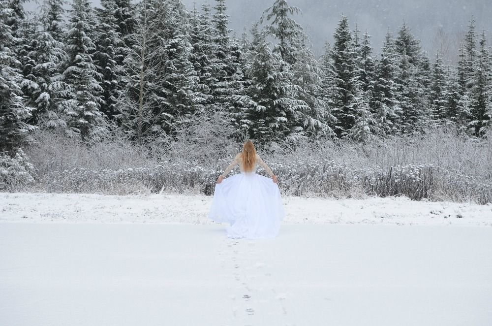Dr. Michele Burklund  in a white dress walking through snow in a winter landscape with snow-covered trees in the background.