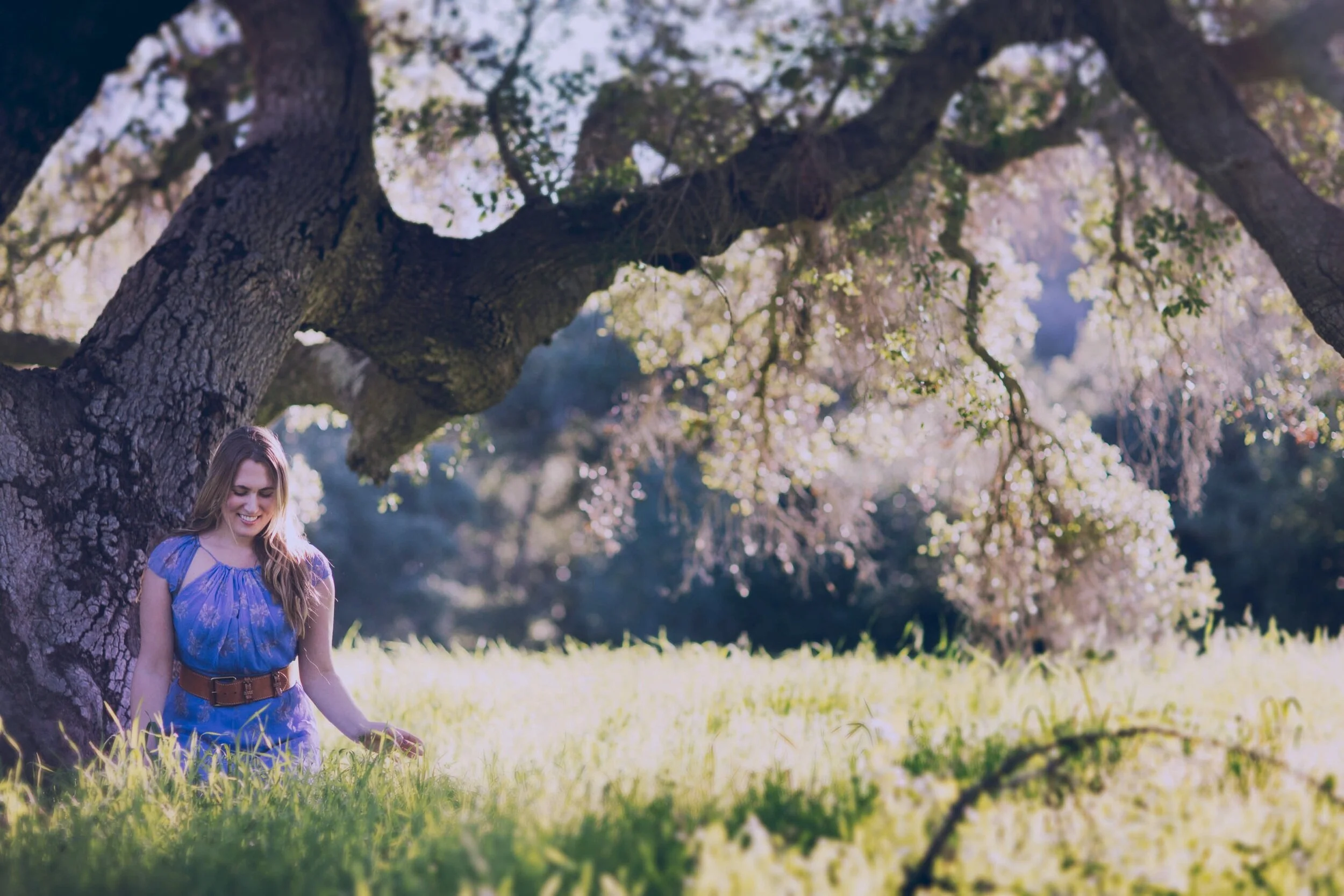 Dr. Michele Burklund  in a blue dress sitting on the grass next to a large tree in a park, surrounded by sunlight and blossoming trees.