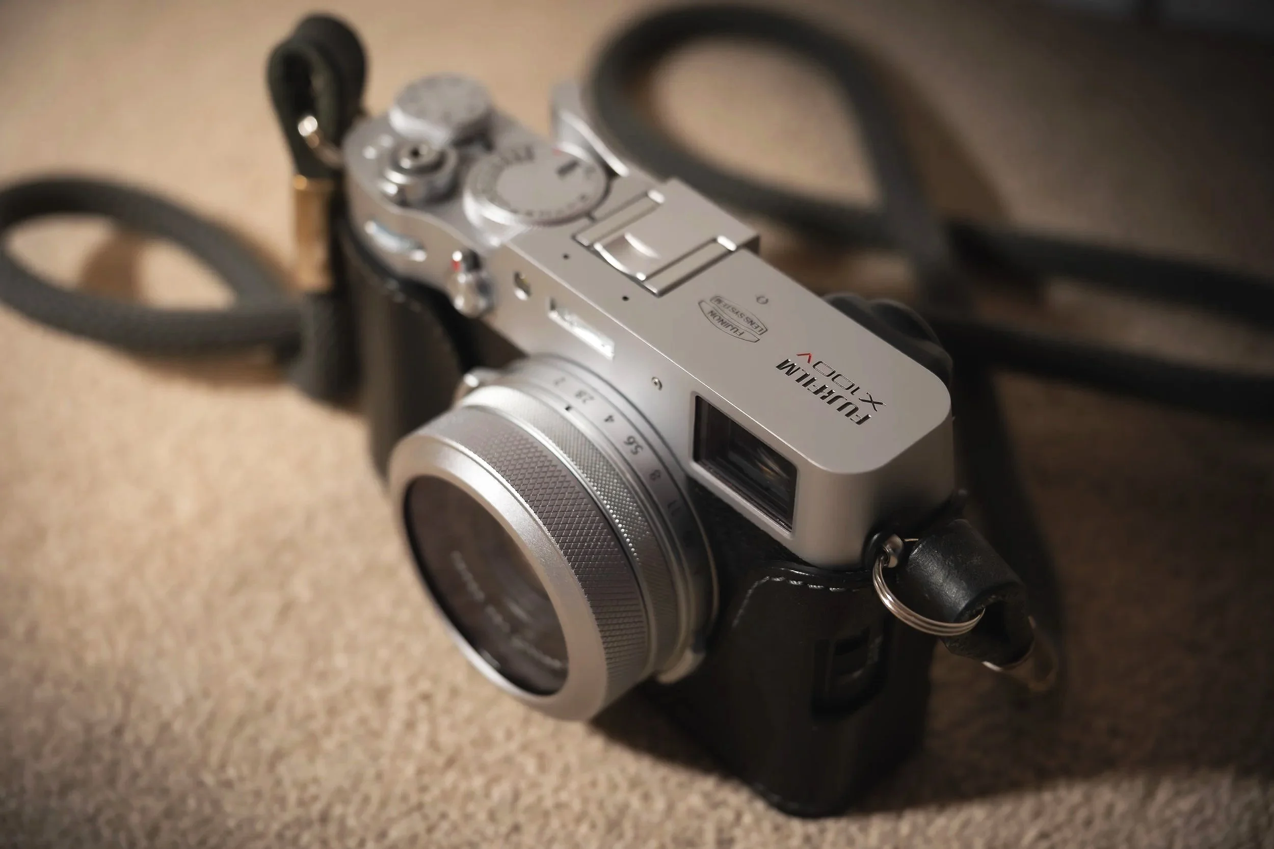 A silver and black Fujifilm X100V camera lying on a textured surface with the strap partially coiled around it.