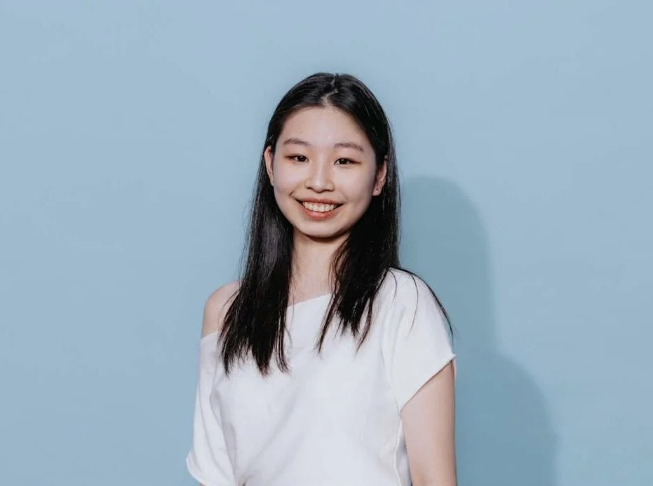 A young woman with long black hair smiling, wearing a white off-shoulder top, standing against a light blue background.