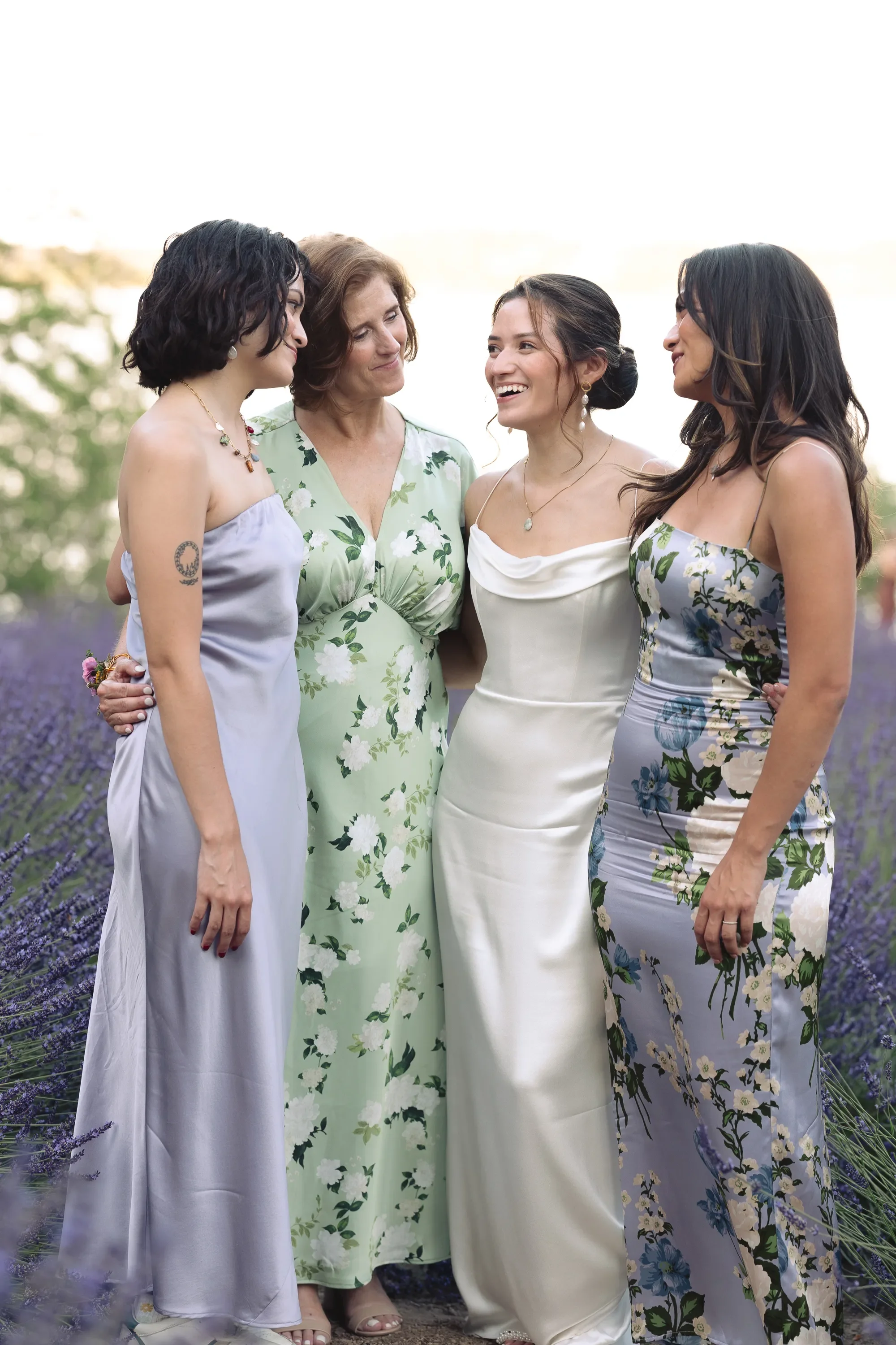 A romantic bridal portrait captured in a lavender field at golden hour, featuring the bride surrounded by her bridesmaids in soft, floral, and pastel dresses. This timeless group moment highlights natural emotion, connection, and elegant styling, per