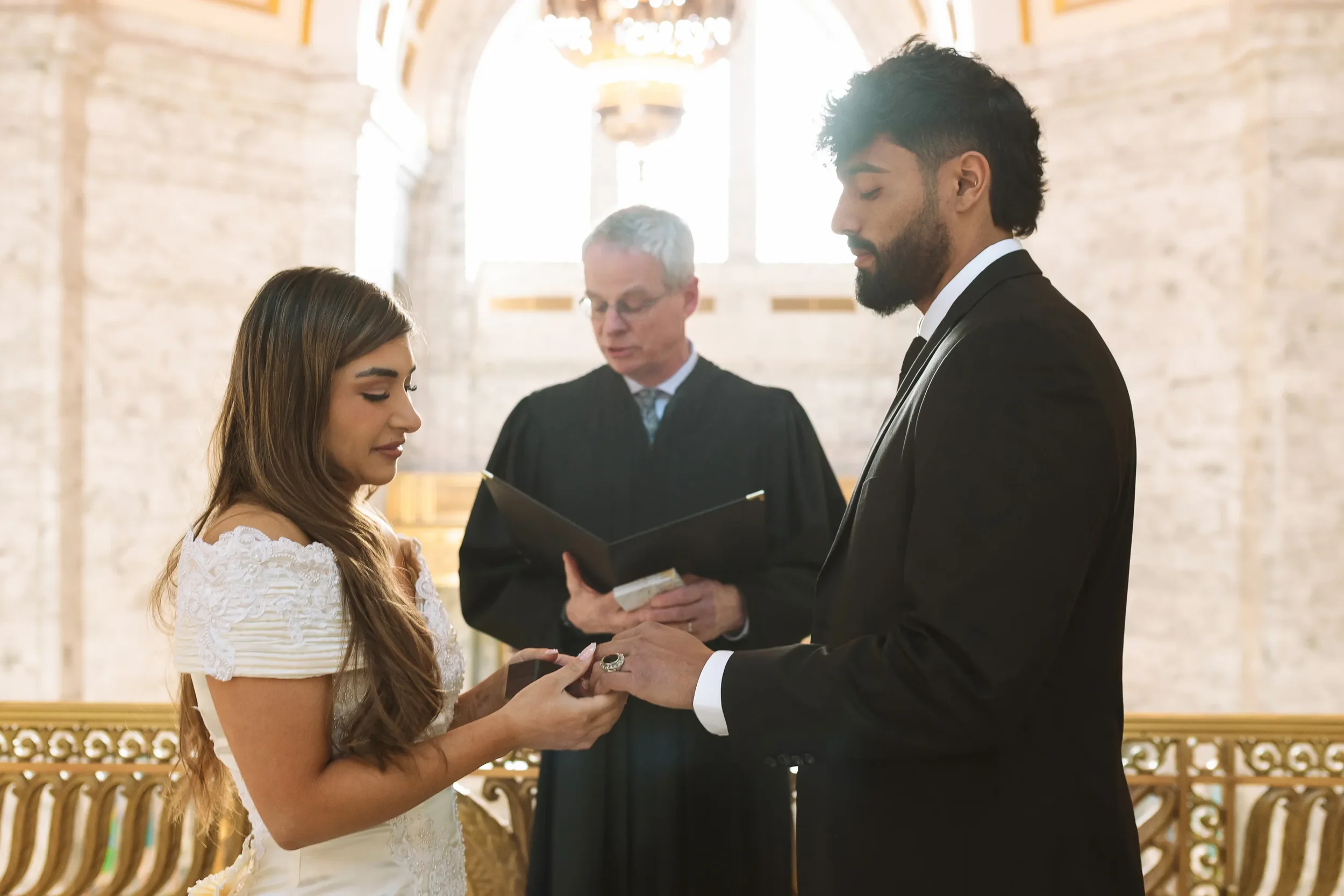 A bride places a wedding ring on her partner’s finger during an intimate civil ceremony inside the Washington State Capitol, with an officiant standing before marble walls and arched windows.