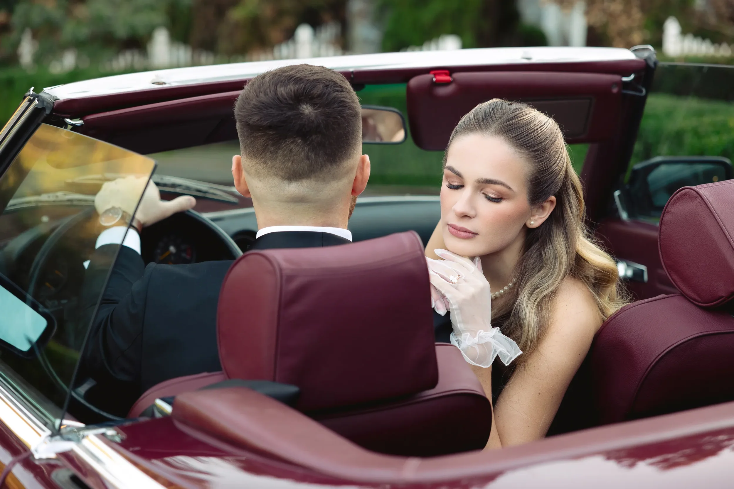 A couple sitting in a vintage convertible with maroon leather seats during their Fox Hollow Farm wedding, featuring the groom in a dark suit and the bride in elegant sheer gloves and pearls, captured in a candid, editorial moment.