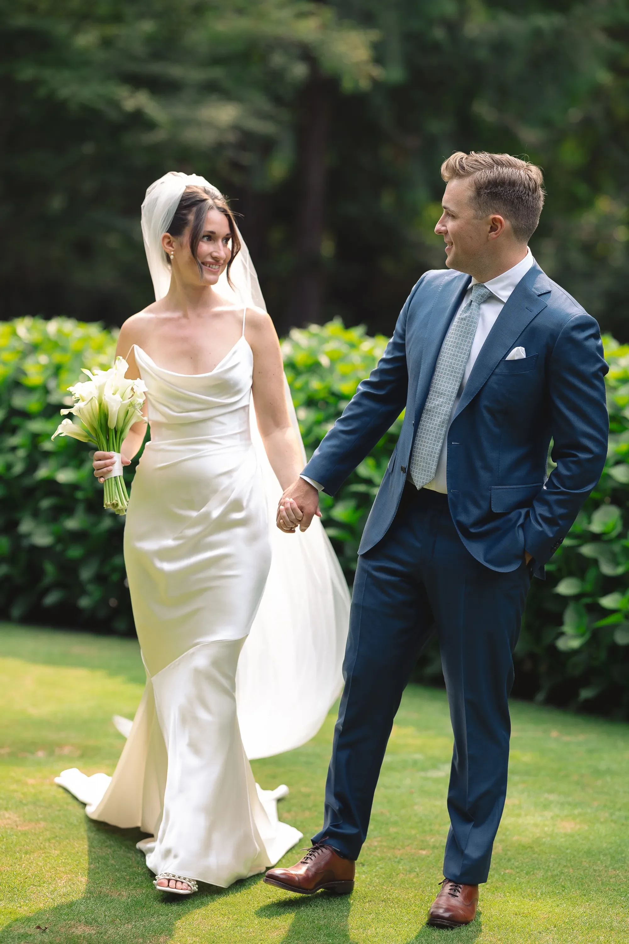 A romantic wedding portrait of a bride and groom walking hand in hand through a lush garden, captured in natural light. The bride wears a minimalist silk gown with a soft draped neckline and veil, holding a bouquet of white calla lilies, while the gr