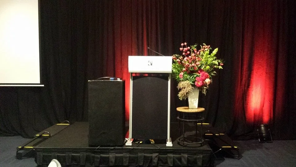 Vibrant Corporate speaker lectern arrangement in reds and green, featuring fresh Kale, Dahlia, Irish Bells, Ivy Berry, and artificial Chinese Lanterns.