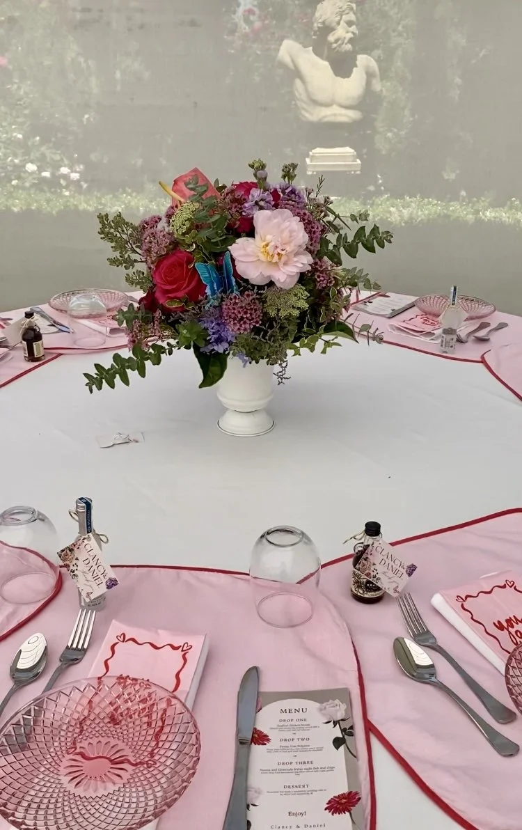 Colourful spring wedding guest table centrepiece in white metal urn, featuring pink peonies, not pink roses, lilac stocks, purple misty, wax flower, queen anne’s lace, and fresh greenery. Styled with blush pink linen and red-edged napkins. Masonmill