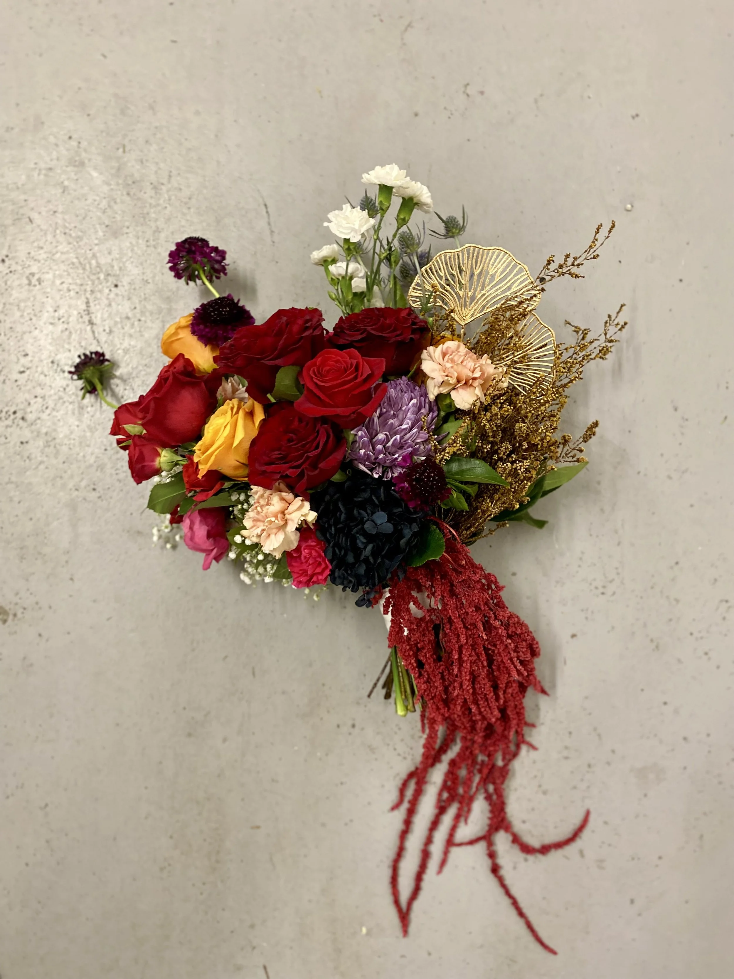 Modern wedding bouquet featuring Red, burgundy and mustard-gold roses, burgundy scabiosa, purple disbud, peach carnation, white spray carnation gold ginko and hanging amaranthus preserved. Designed and created by Perth wedding florist Penny Dormer.