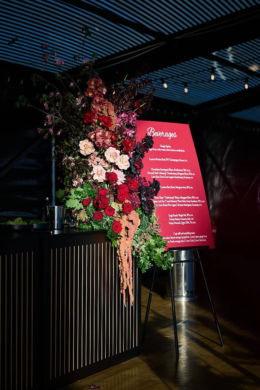 Lush statement bar floral installation in deep red, burgundy, and blush tones with trailing amaranthus, roses, cymbidium orchids and peonies designed for a moody event setting. Designed and created by Penny Dormer of Wild Heart Flowers – Perth Event 