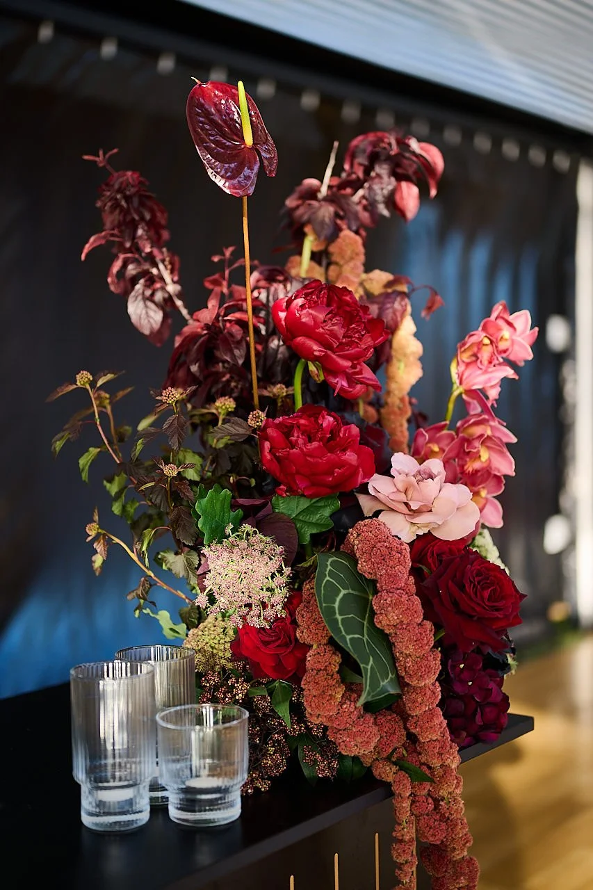 Artistic registration desk floral arrangement in layered burgundy, pink and blush blooms with cascading texture and deep foliage, styled in a modern black setting. Designed and created by Penny Dormer of Wild Heart Flowers – Perth Event Florist.