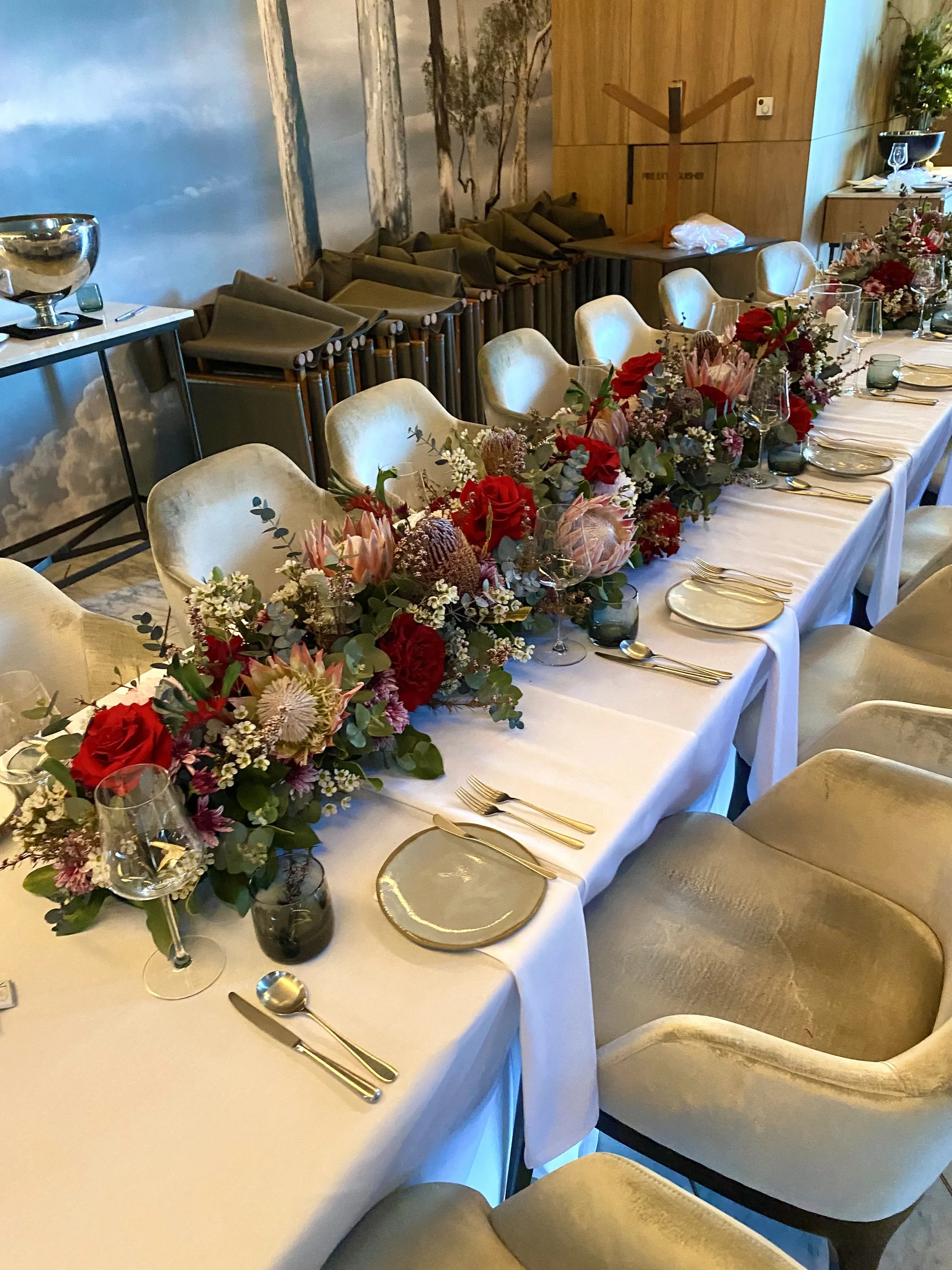 Close-up angled view of wedding table flowers at Wildflower Restaurant, with long garland arrangements of proteas, banksia, red roses, pink wax flower, and gum leaves. Modern Australian native wedding flowers by Penny Dormer, Perth wedding florist.