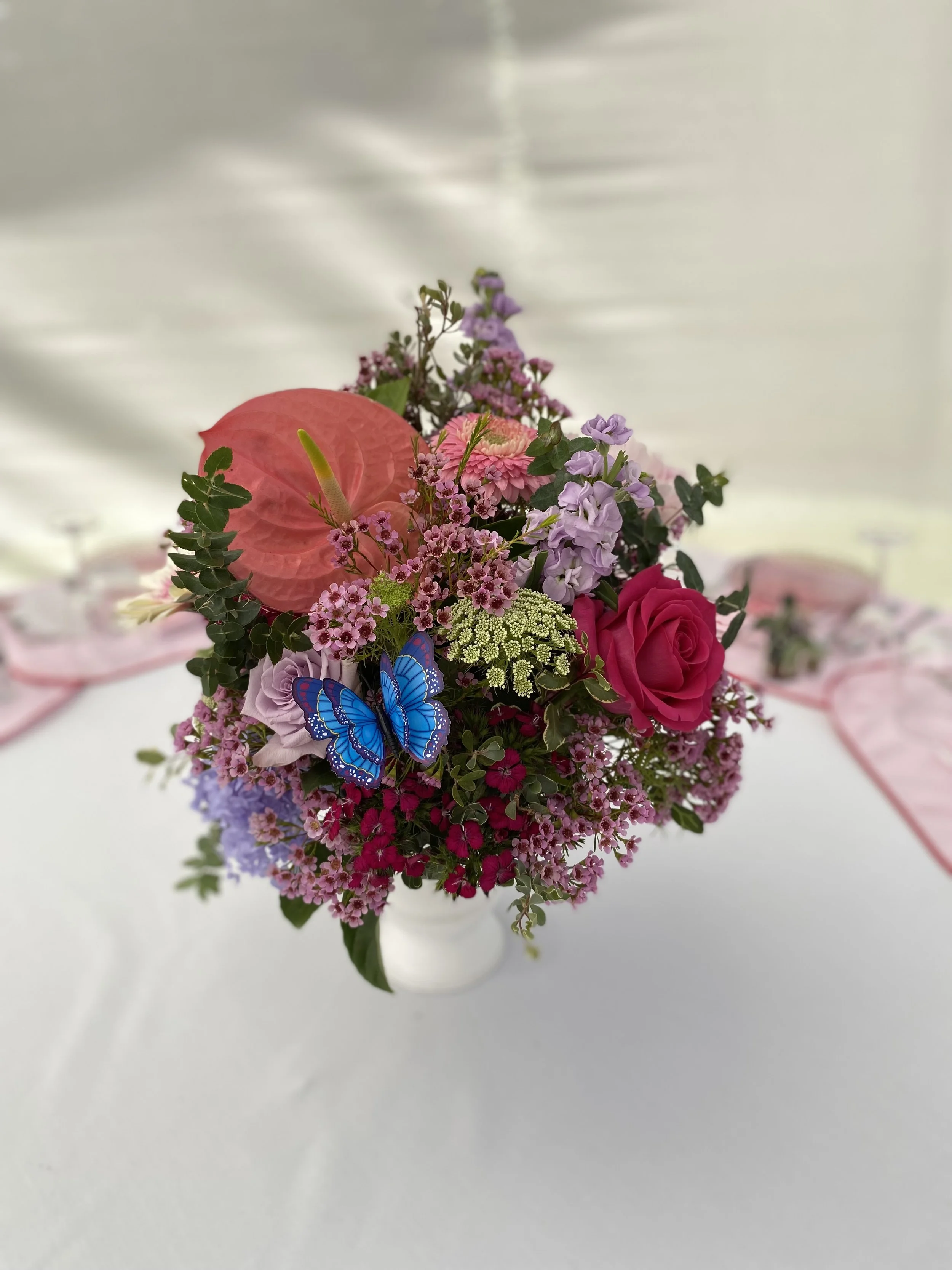 Colourful wedding table centrepiece with bright pink roses, coral anthurium, lavender lisianthus, wax flower, and green queen anne’s lace. Accented with a decorative blue butterfly. Spring wedding florals at Masonmill Gardens by Penny Dormer.