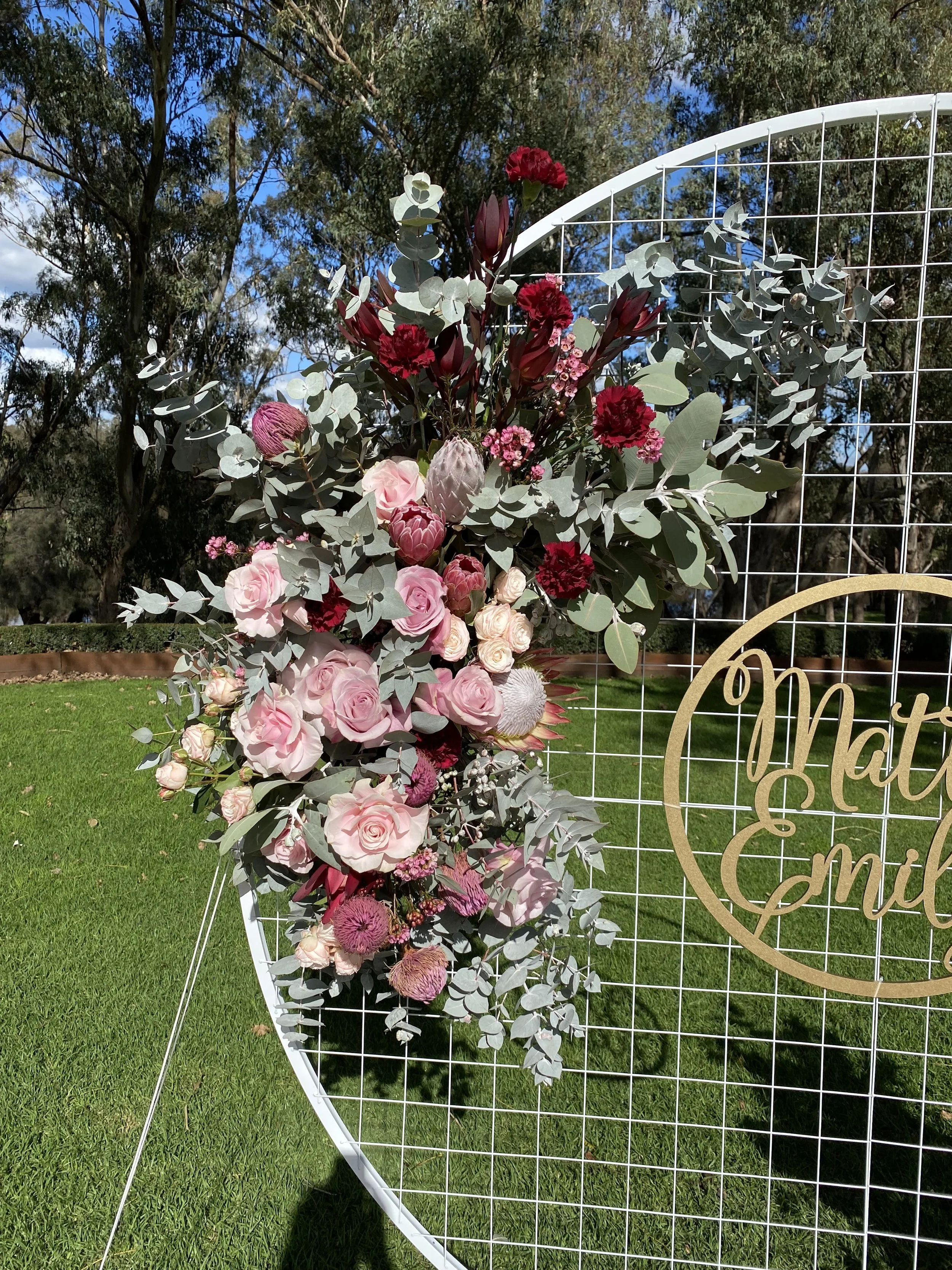 Close up of side floral piece of an outdoor wedding ceremony backdrop featuring a round white mesh frame with floral clusters in blush pink, soft mauve, and deep burgundy tones. Flowers include roses, leucadendron, carnations, and eucalyptus foliage.