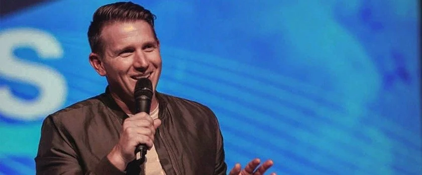 A man smiling and speaking into a microphone on stage with a blue background.