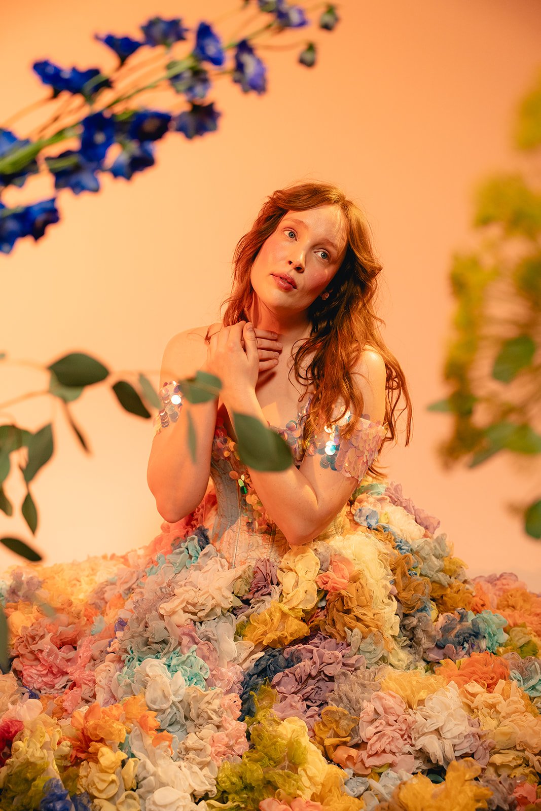 A woman with red hair, wearing a dress made of colorful fabric flowers, is surrounded by more fabric flowers and greenery, with a warm orange background - portrait photographer Cork