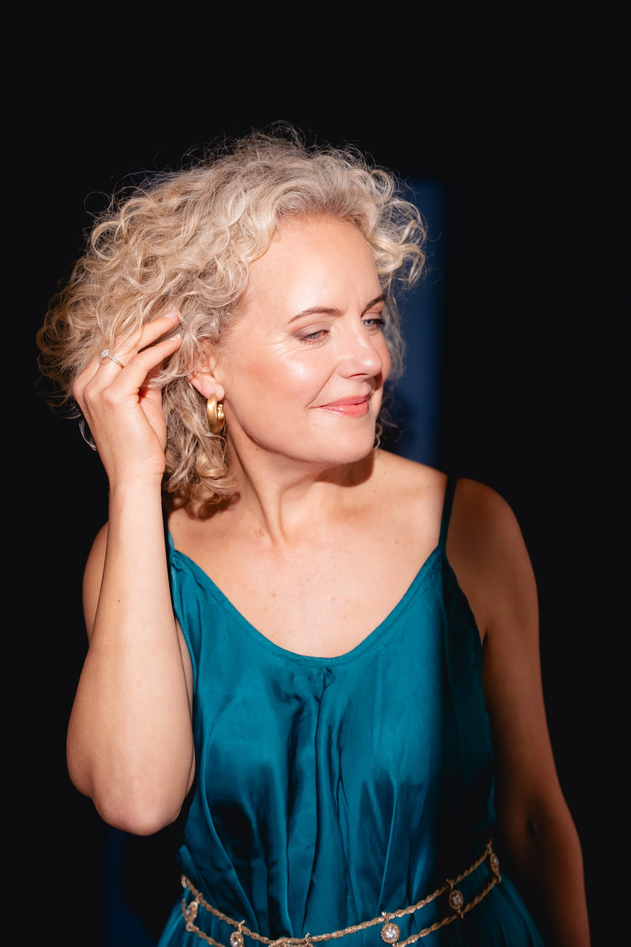A blonde woman puts her hands in her hair. The spotlight lights her up in a Cork photo studio. She wears a dark blue dress and looks away from the camera. Professional headshots Cork Ireland.