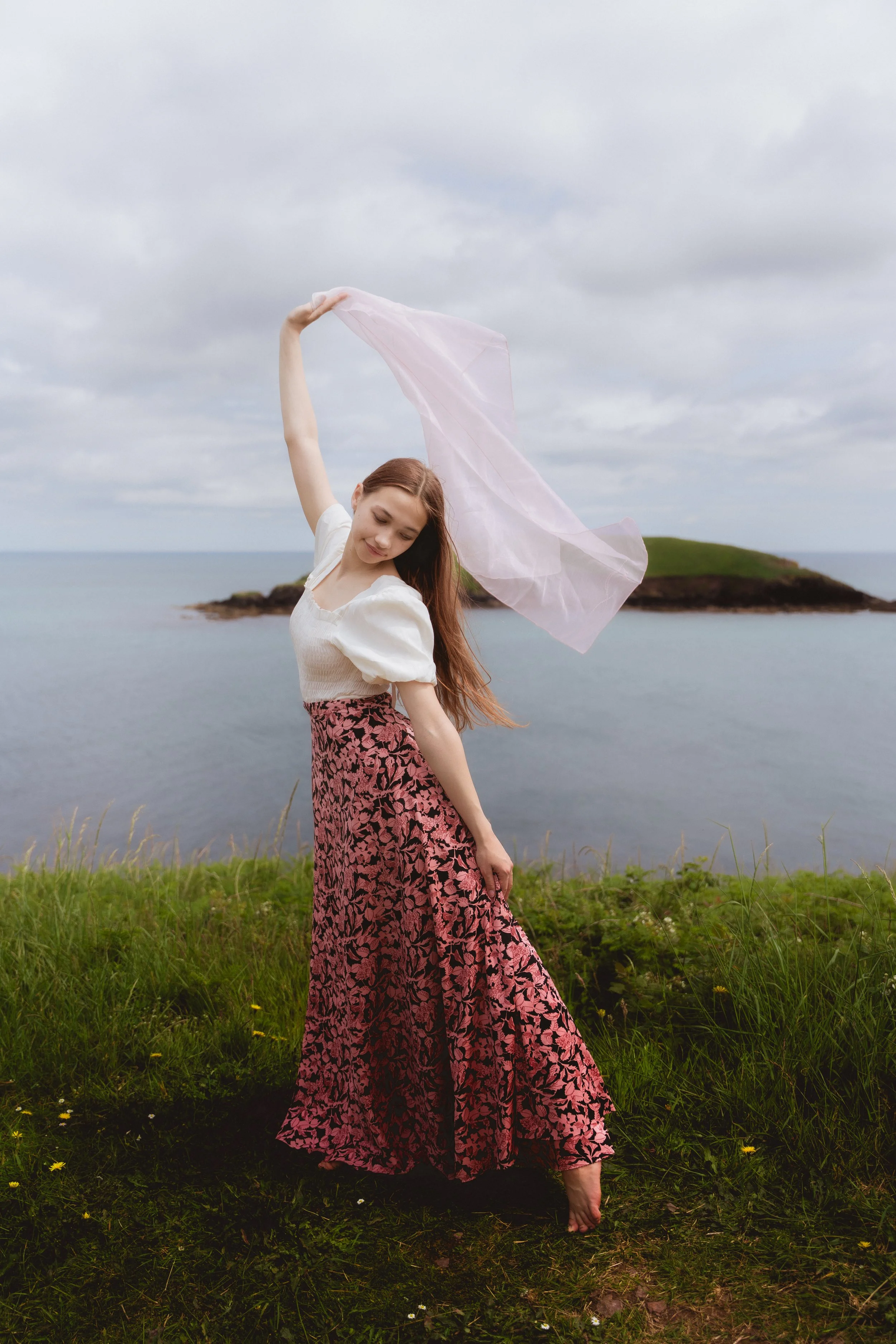 A young woman in a pink dress holds a pink piece of fabric that moves because of the wind. She stand bare-feet on the grass near the sea. Behind her we see a small island in Knockadoon. Portrait photographer Cork.