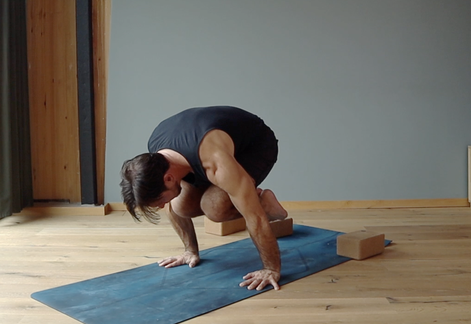 A man is practicing yoga indoors on a blue yoga mat, balancing on his hands and feet with his knees bent, using yoga blocks for support.