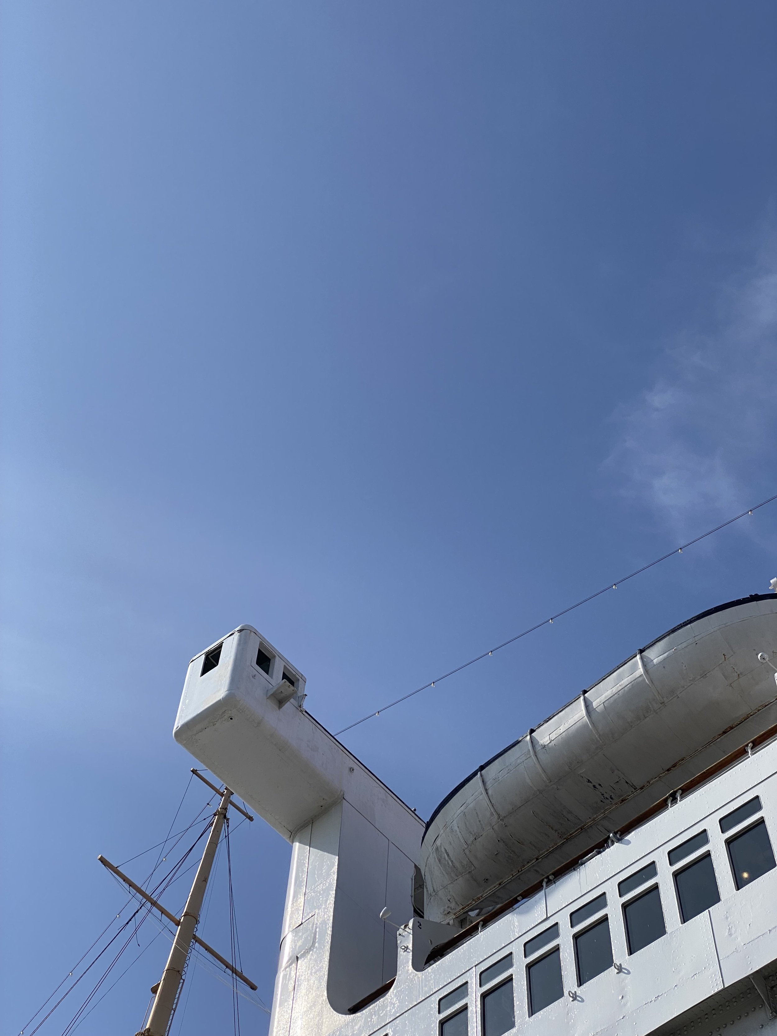 Close-up of the upper part of a white ship with an observation post, a mast, festoon lights and the underneath of lifeboat, against a clear blue sky.