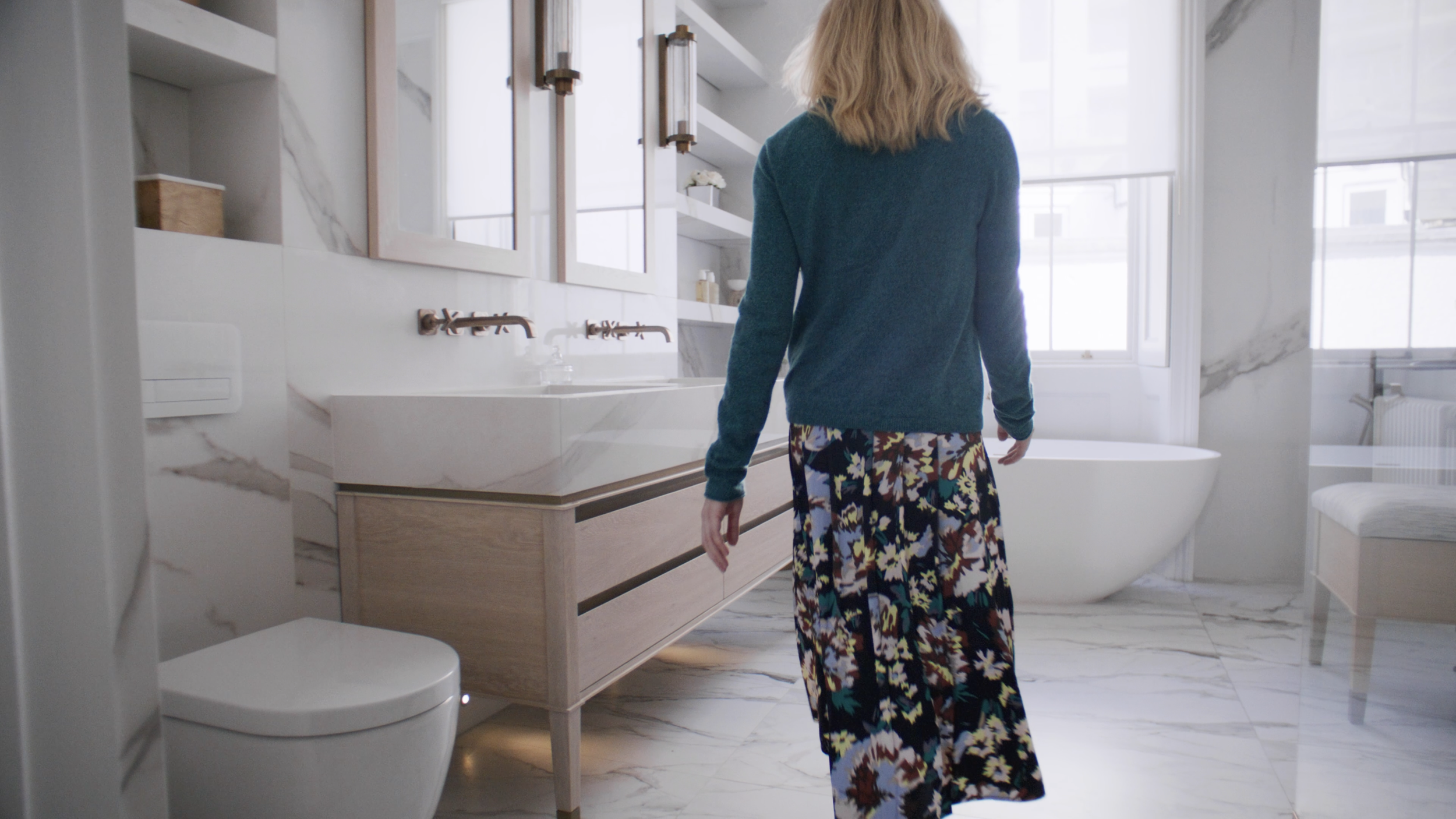 A woman with blonde hair walking in a modern bathroom with white marble walls and floor, a white bathtub near the window, and a wooden vanity with a marble top.