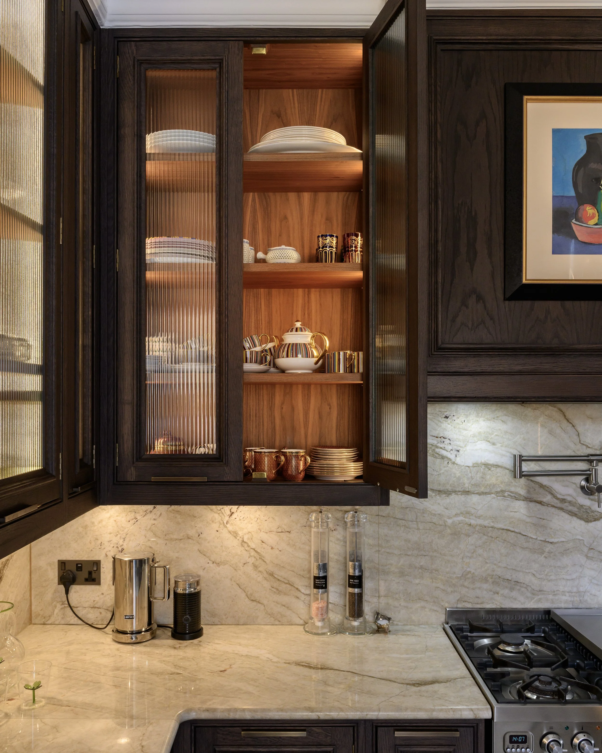A woman with curly blonde hair in a beige dress standing in a modern kitchen, reaching into a refrigerator.