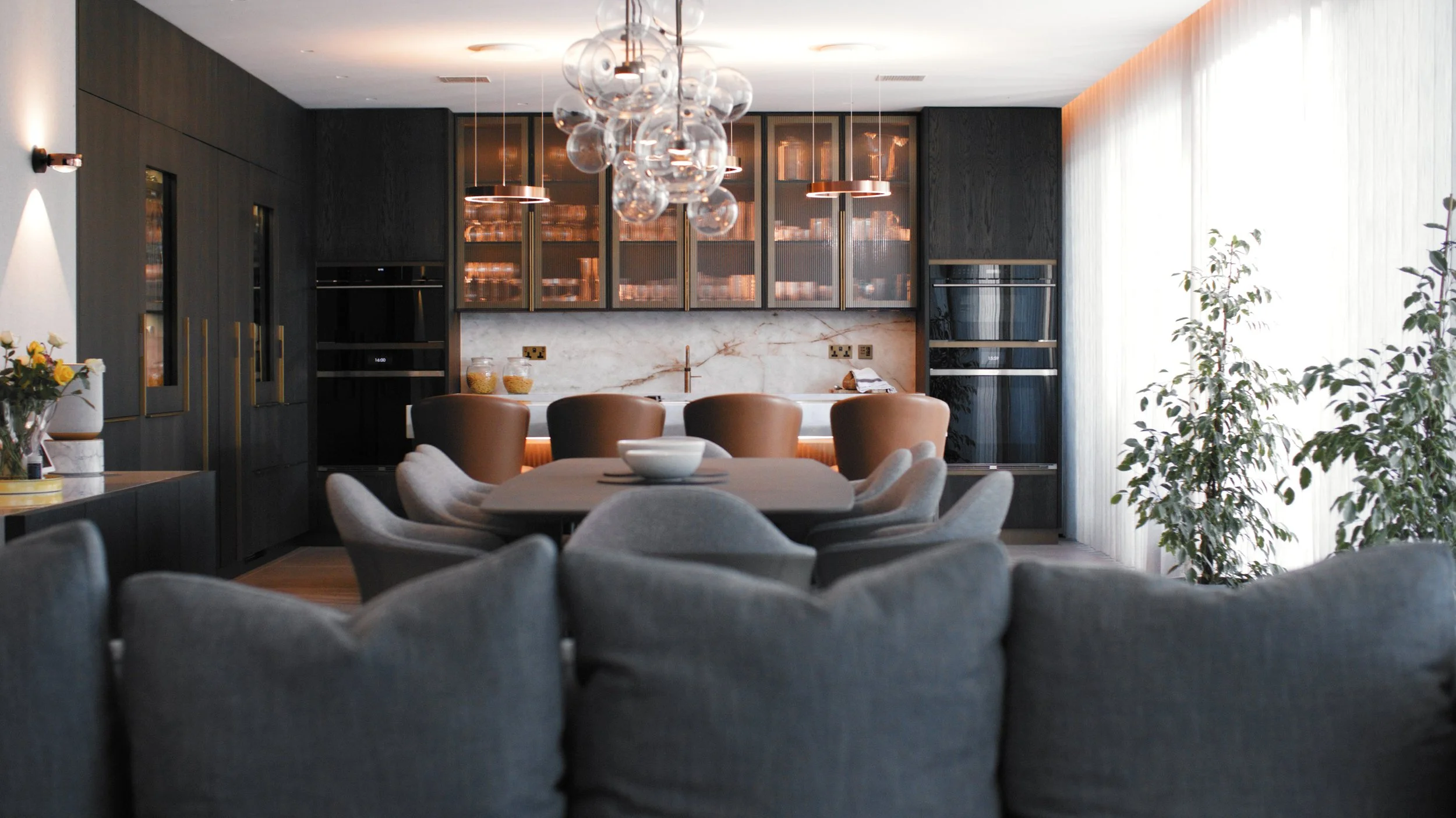 Modern dining room with a round table, gray upholstered chairs, and a chandelier with glass globes, featuring dark wood cabinetry and large windows with sheer curtains.