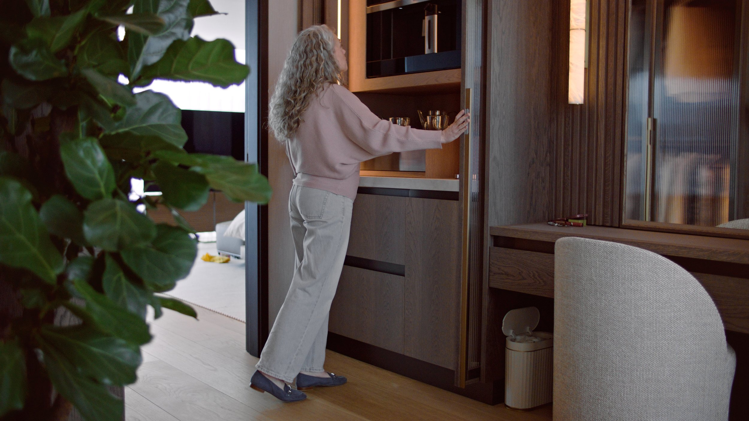 A woman with curly blonde hair holding a glass with a drink in a wooden cabinet in a modern living space.