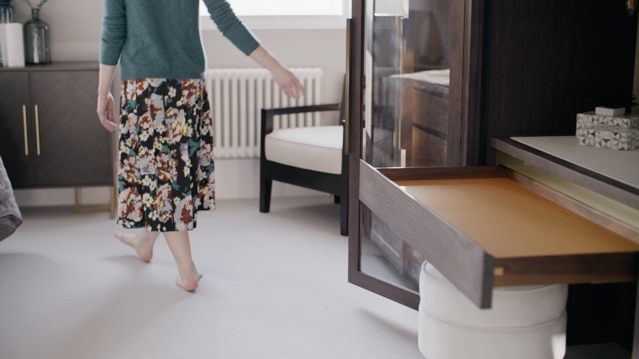 A person is walking barefoot in a bedroom, wearing a teal sweater and floral-patterned pants. There is a desk with a pull-out tray, a chair, a white radiator, and various decorative items in the room.