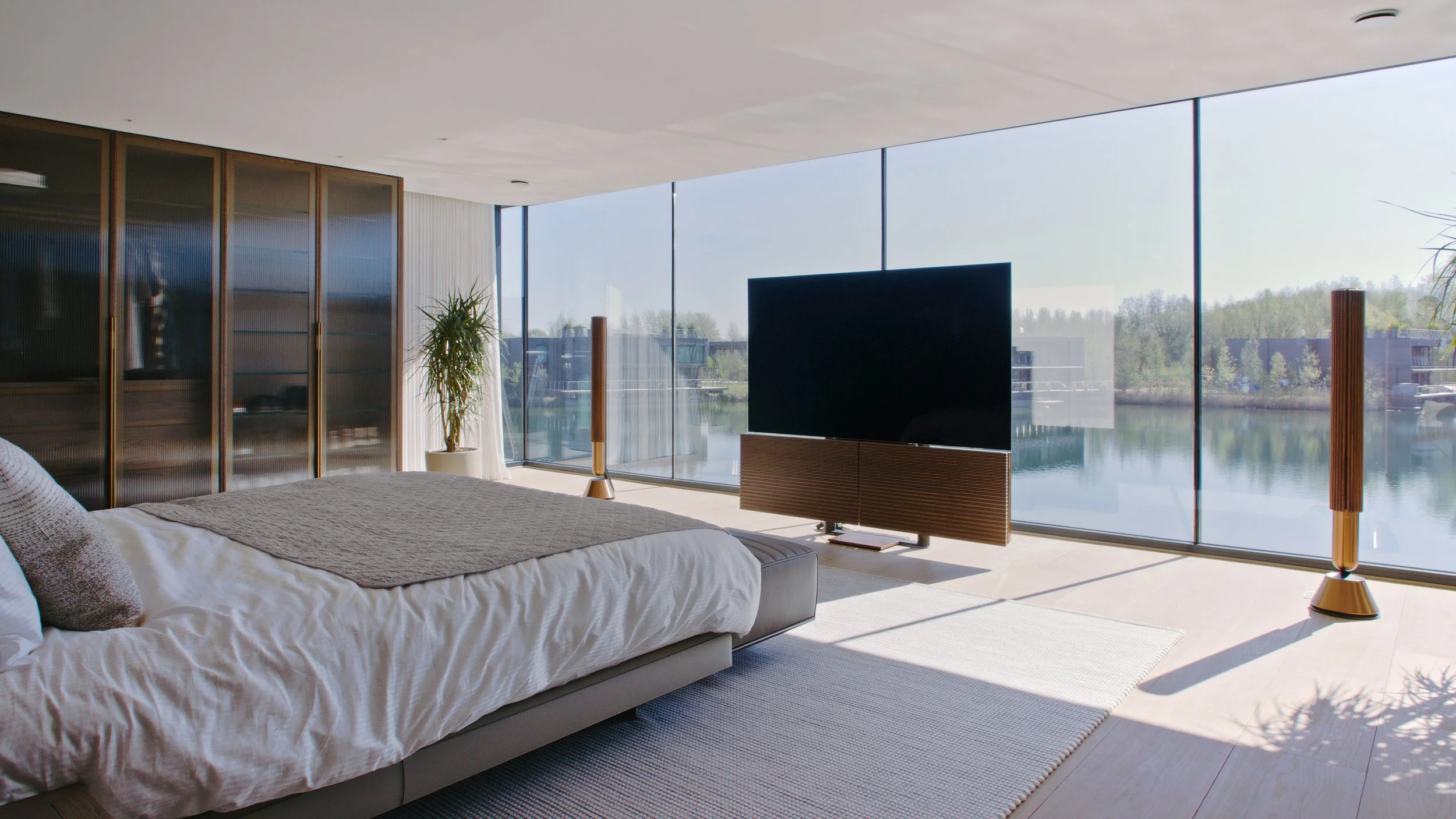 Modern bedroom with a large bed, a flat-screen TV, and floor-to-ceiling windows overlooking a body of water with trees in the distance.